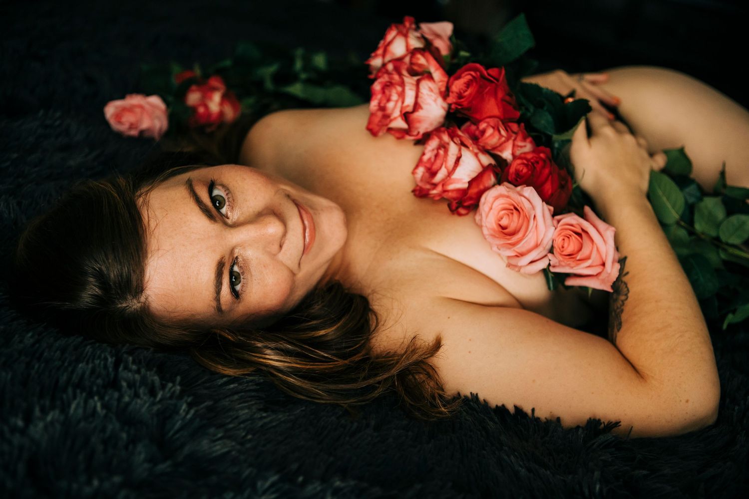 A woman poses with pink roses against a dark backdrop in an artistic boudoir photoshoot.