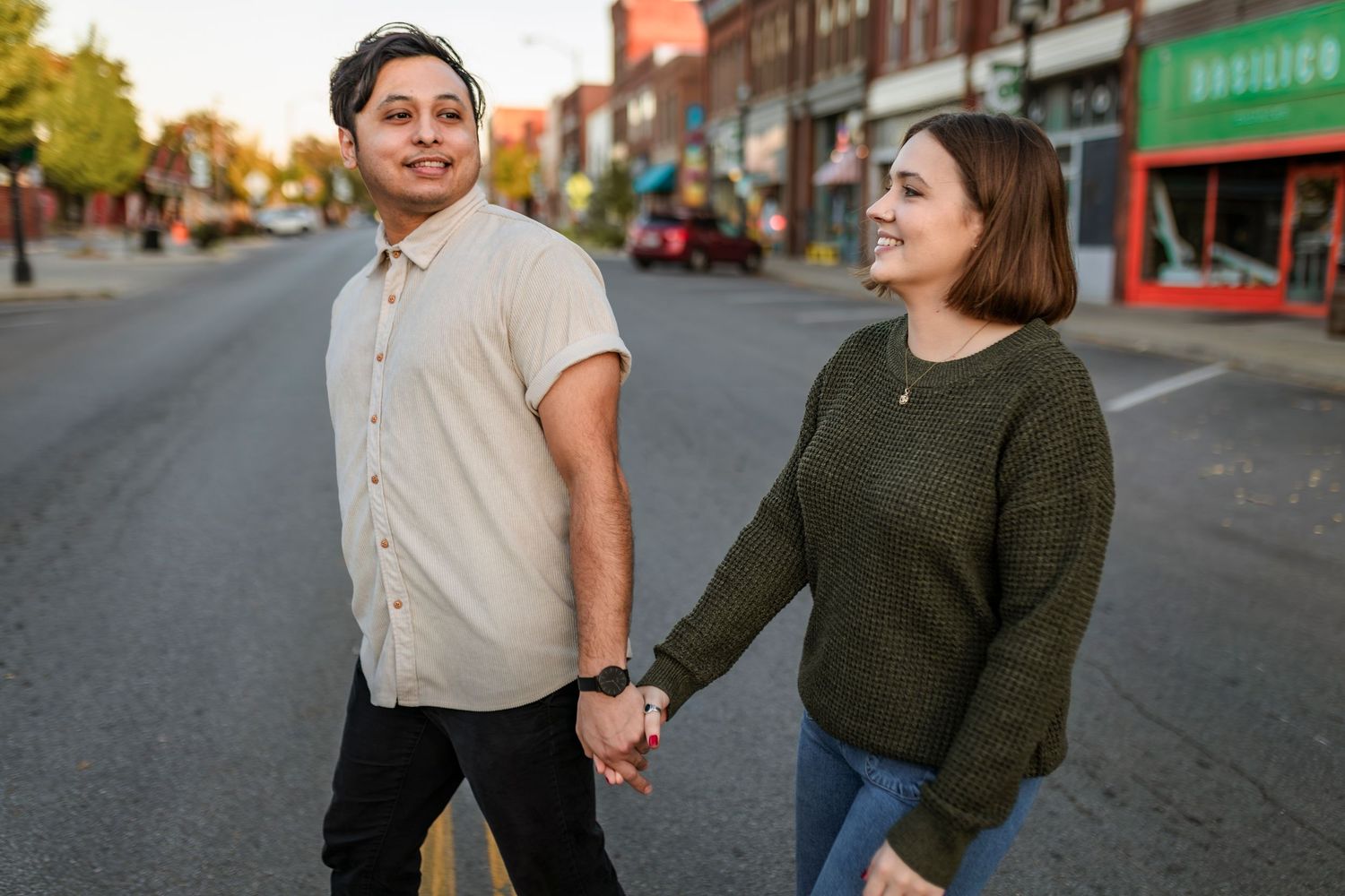 A couple holding hands walks down a city street at golden hour with brick buildings and storefronts in the background.