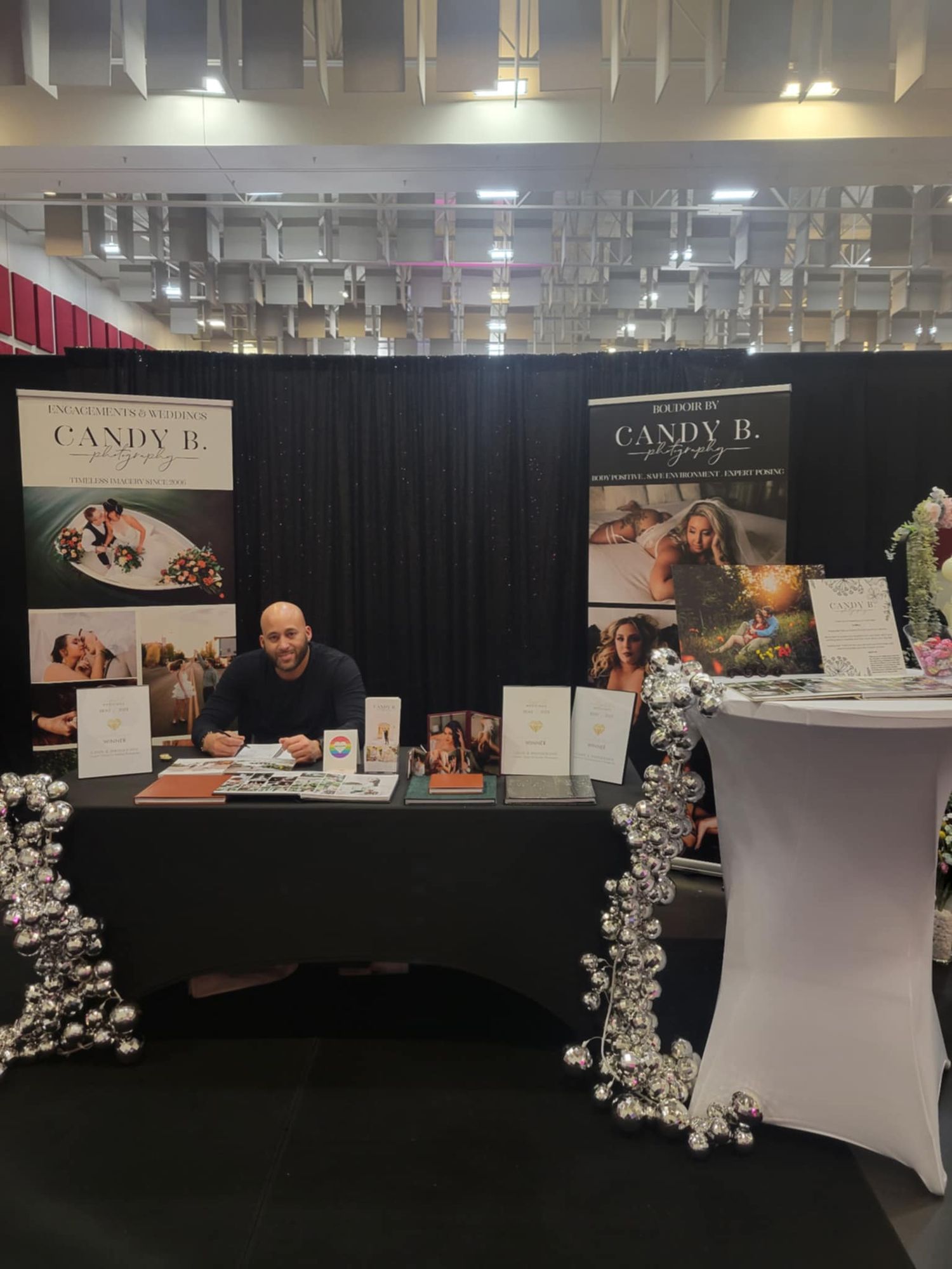 A professional photography studio booth setup at a trade show with black curtains, white tables and floral decorations.