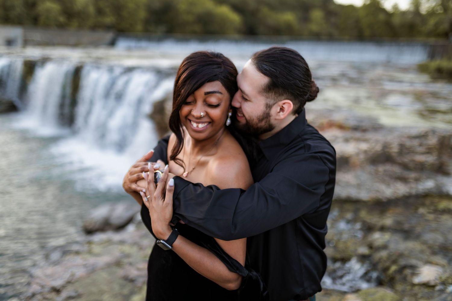 A romantic couple shares an intimate moment together by a waterfall, with one partner embracing the other from behind.