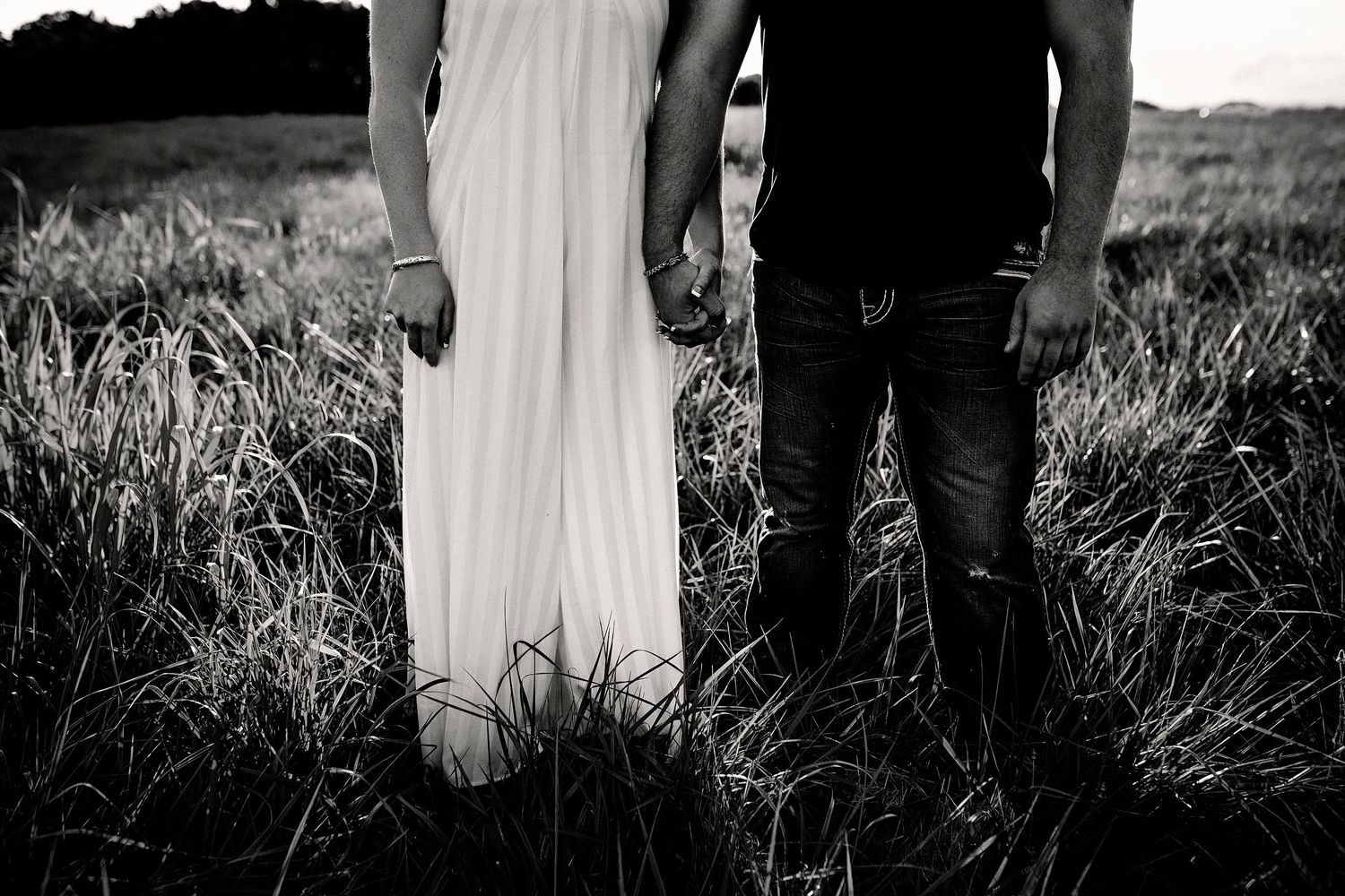 Black and white photo of a couple holding hands while standing in tall grass at sunset on their wedding day.