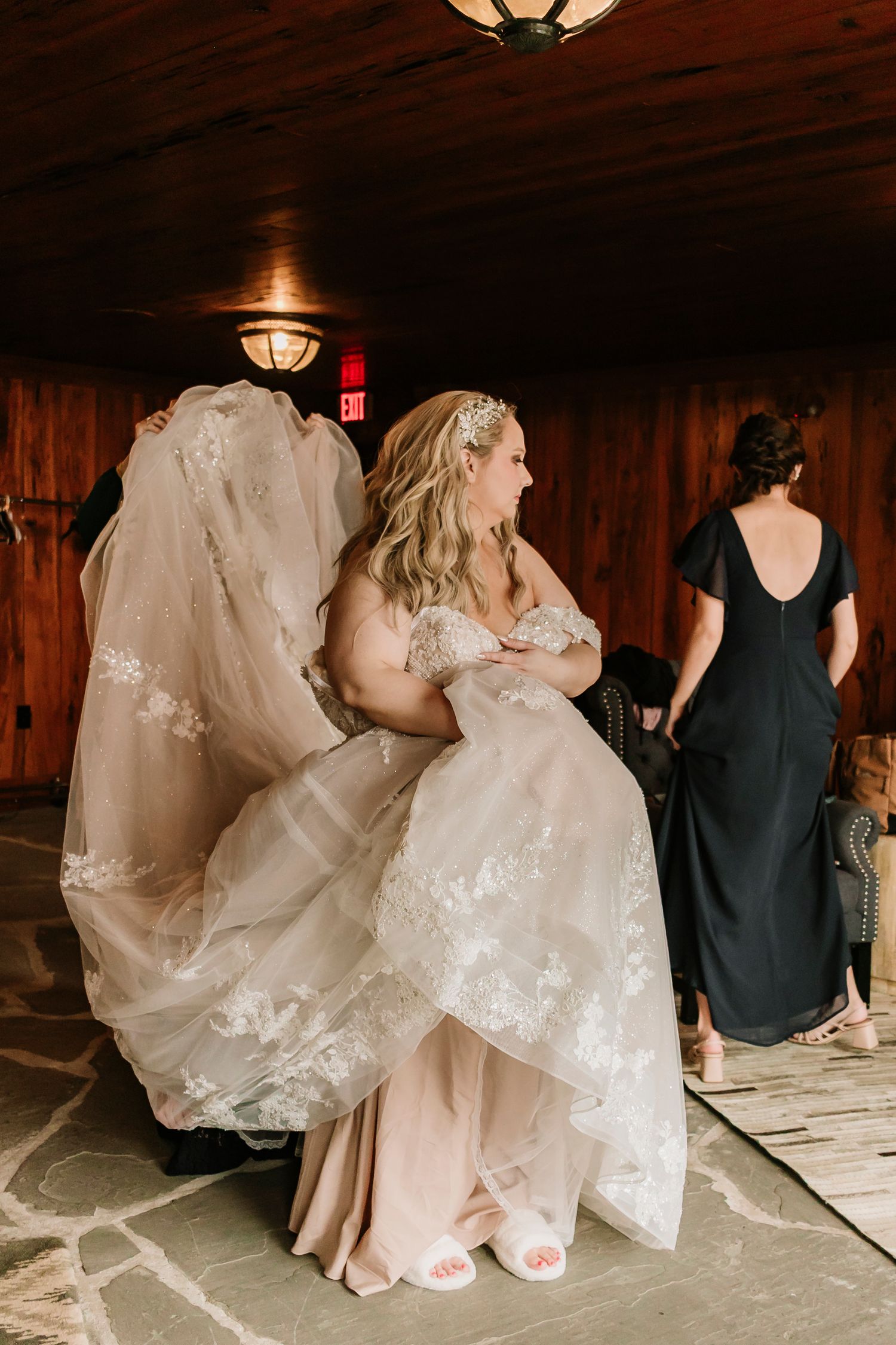 People in wedding attire gather around a wooden bench in a rustic indoor setting.