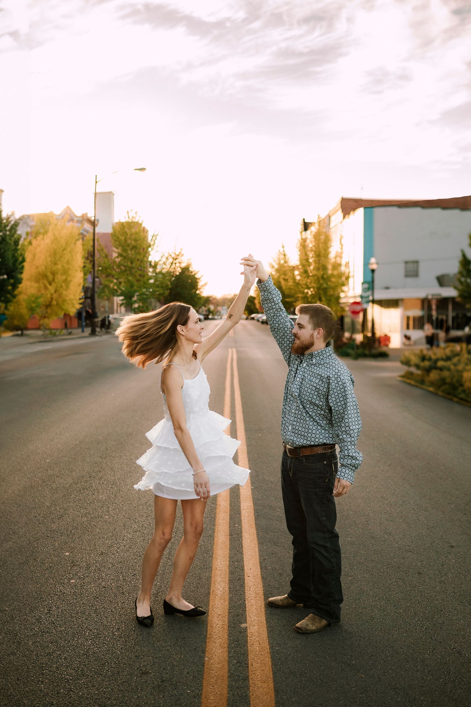 A couple stand together on a street at sunset, with the person in a white dress twirling joyfully.