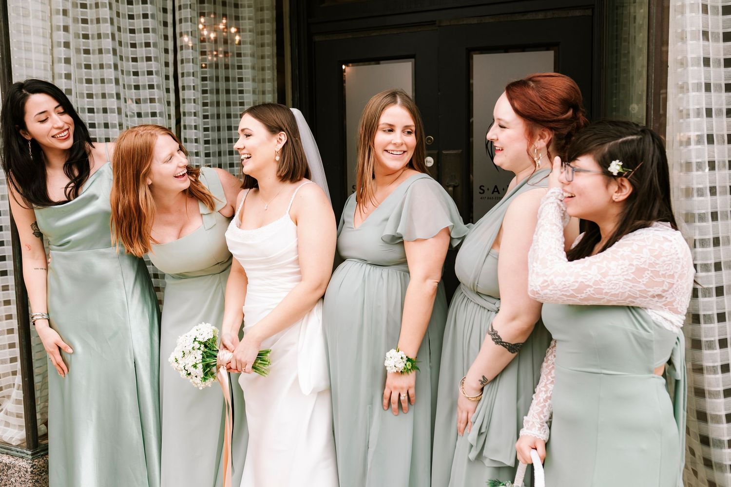 Bridesmaids in sage green dresses smiling and laughing together at a wedding celebration.