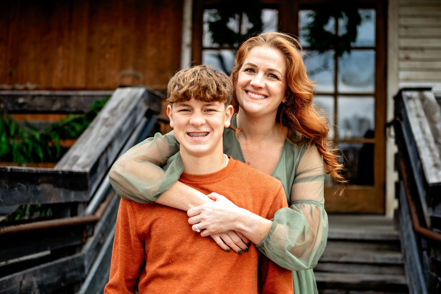Happy parent in sage green cardigan embraces child in orange shirt while standing on rustic wooden porch steps.