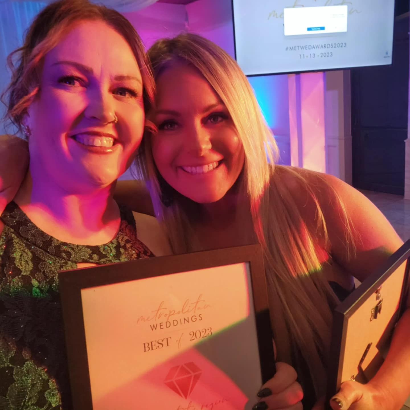 Two people smile while holding award plaques at an event with purple lighting in the background.