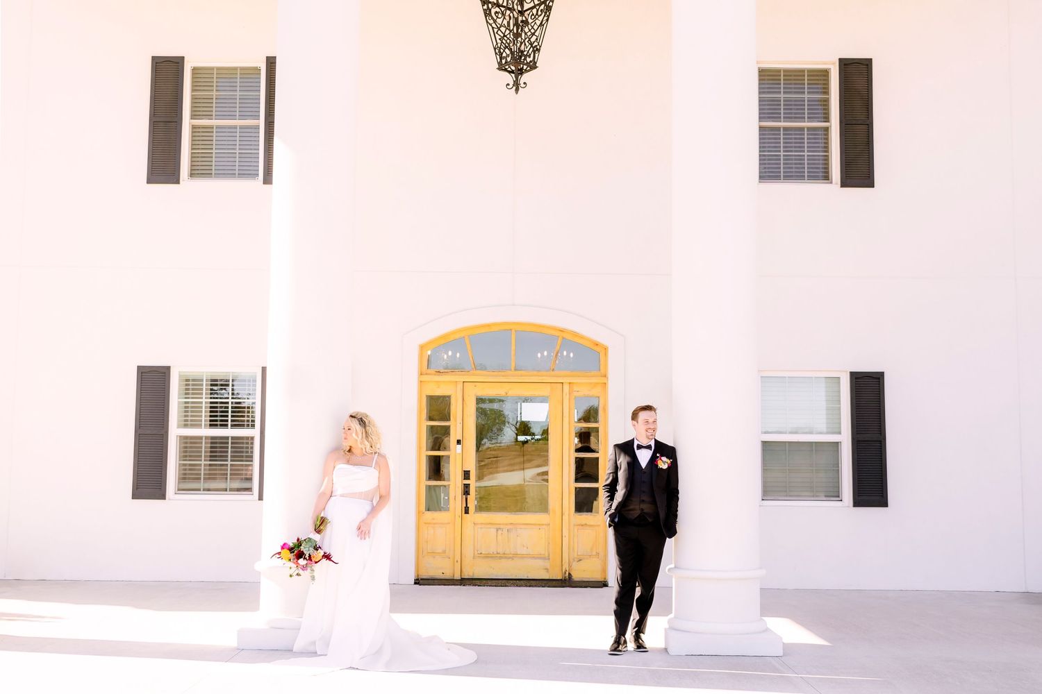 Bride and groom stand separated outside an elegant white building with a gold-framed door and black shuttered windows.
