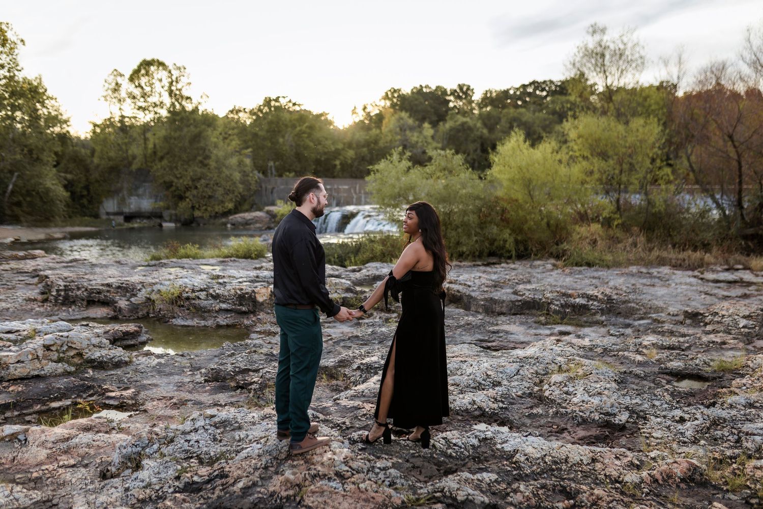 Two people stand facing each other on rocky terrain at sunset with trees and greenery in the background.