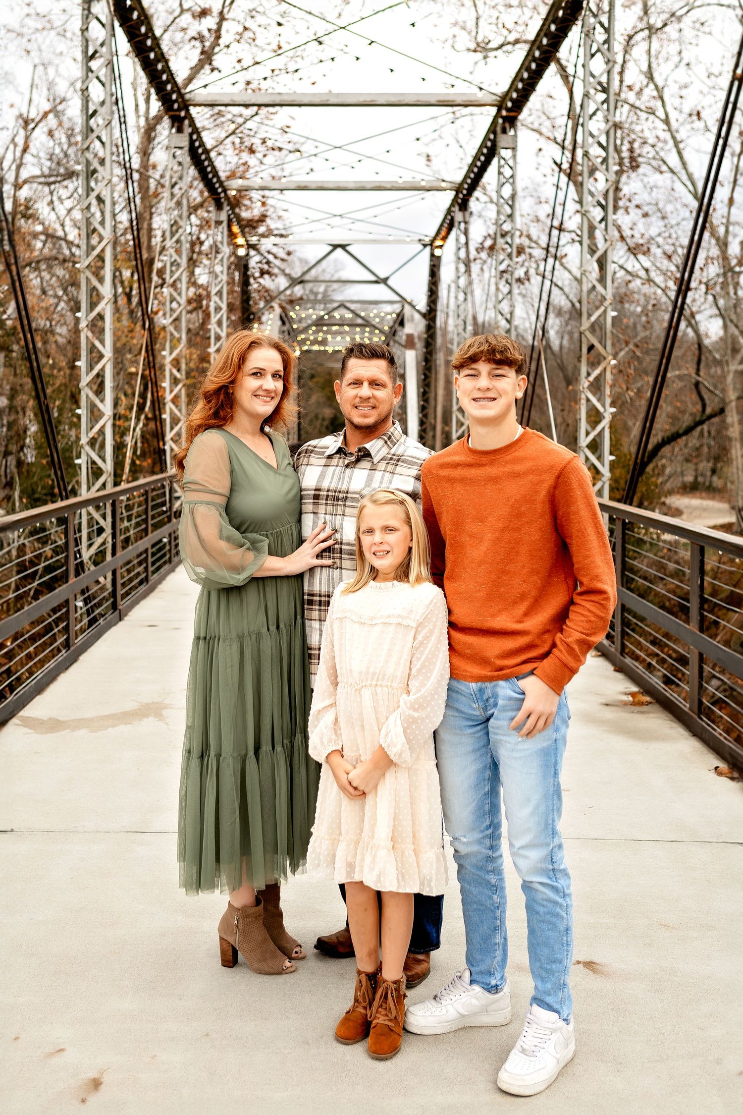 A family stands together on a wooden bridge walkway surrounded by bare winter trees and railings.