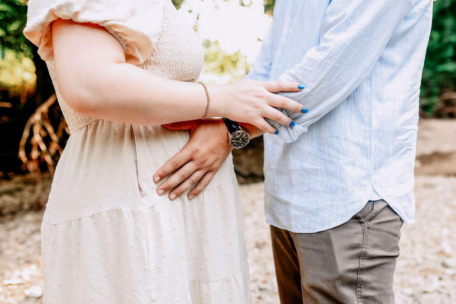 Close-up of an expectant couple holding hands resting on a baby bump, wearing light colored casual clothing outdoors.
