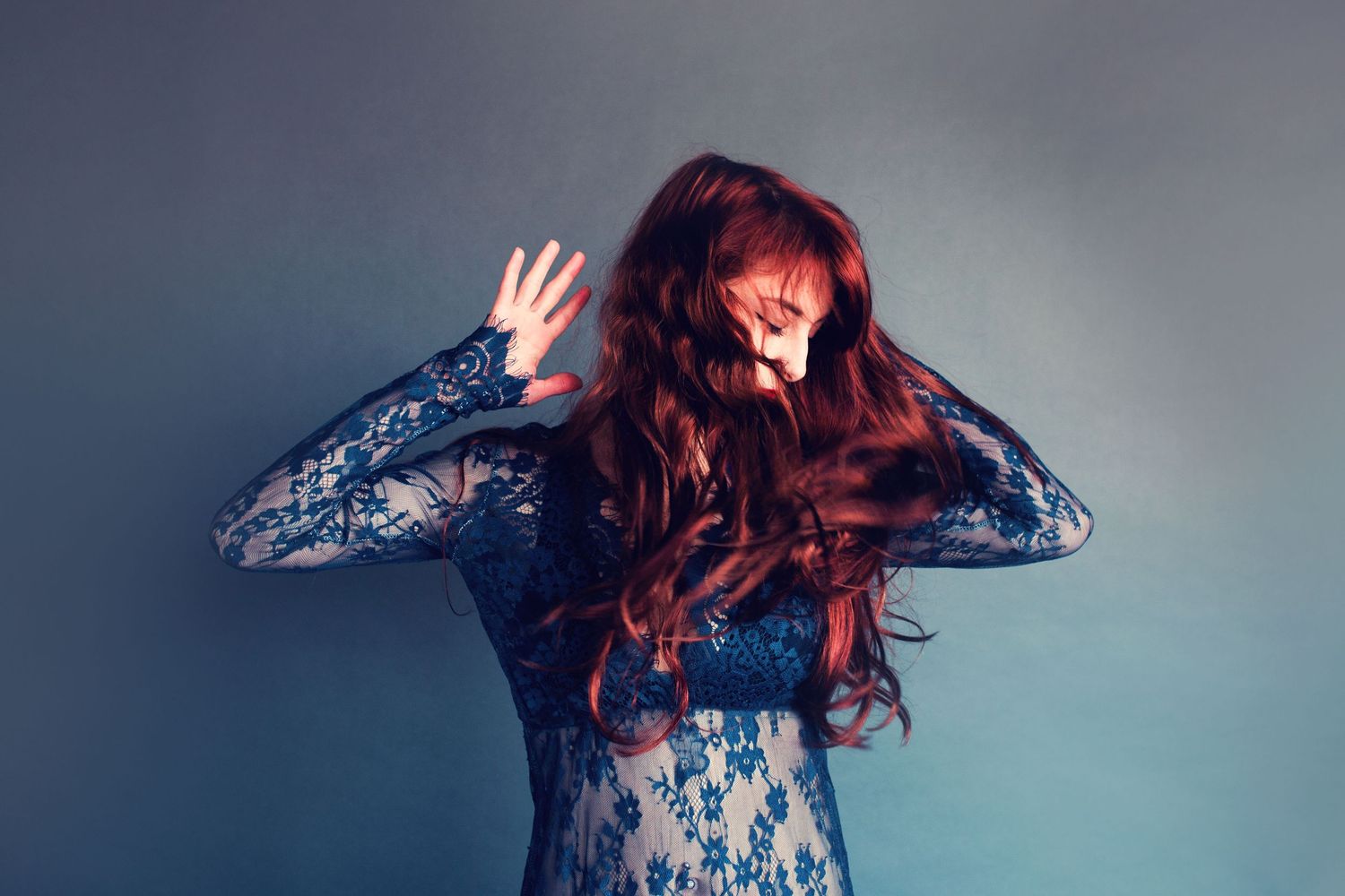A person in a lace dress with long red hair waves their hand in front of their face against a blue-gray background.