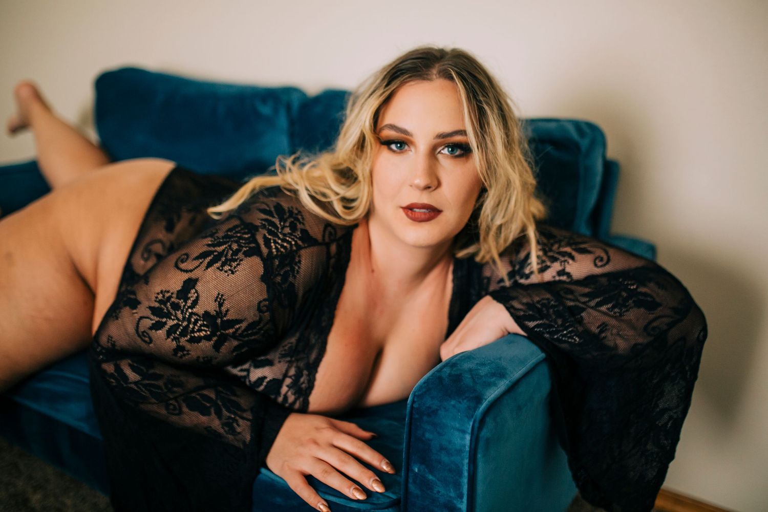 A person wearing a black lace outfit poses elegantly on a blue velvet sofa in a boudoir-style setting.