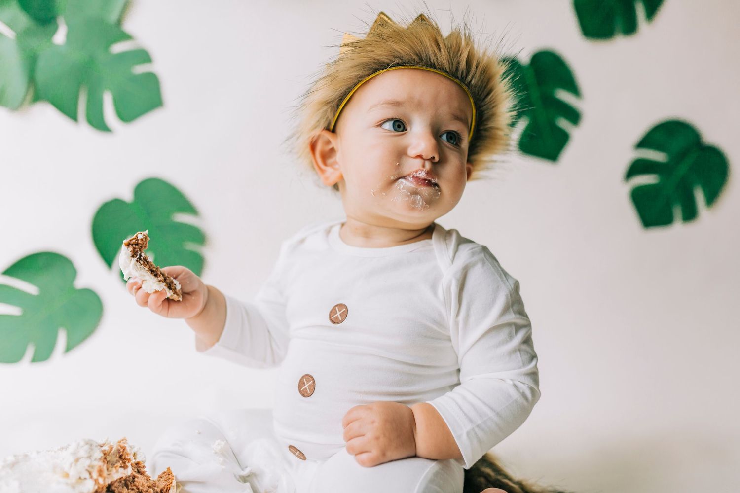 Baby in white button-up shirt sits against white wall with green tropical leaf decorations in background.