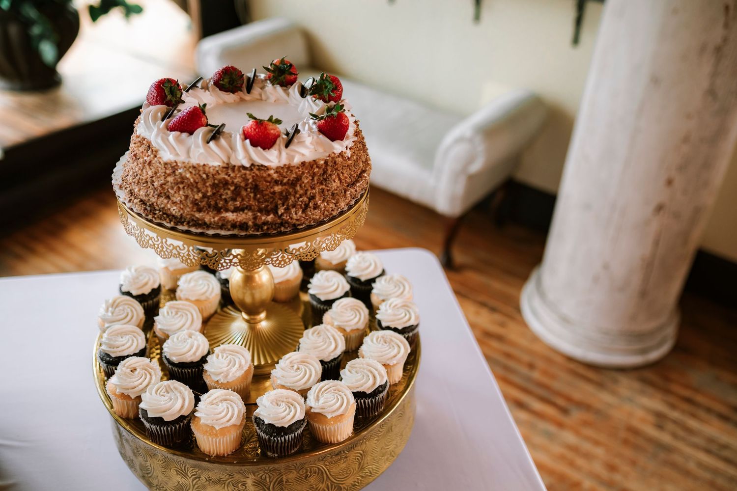 A wedding dessert table displays a decorated chocolate cake with berries and white cupcakes arranged on gold stands.