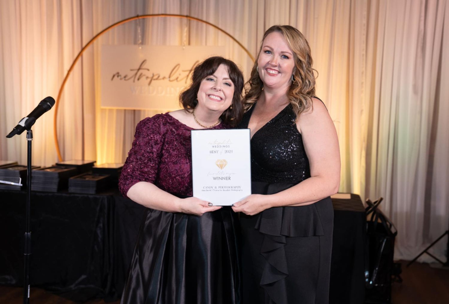 Two performers pose together holding an award certificate in front of a piano and microphone at an event.