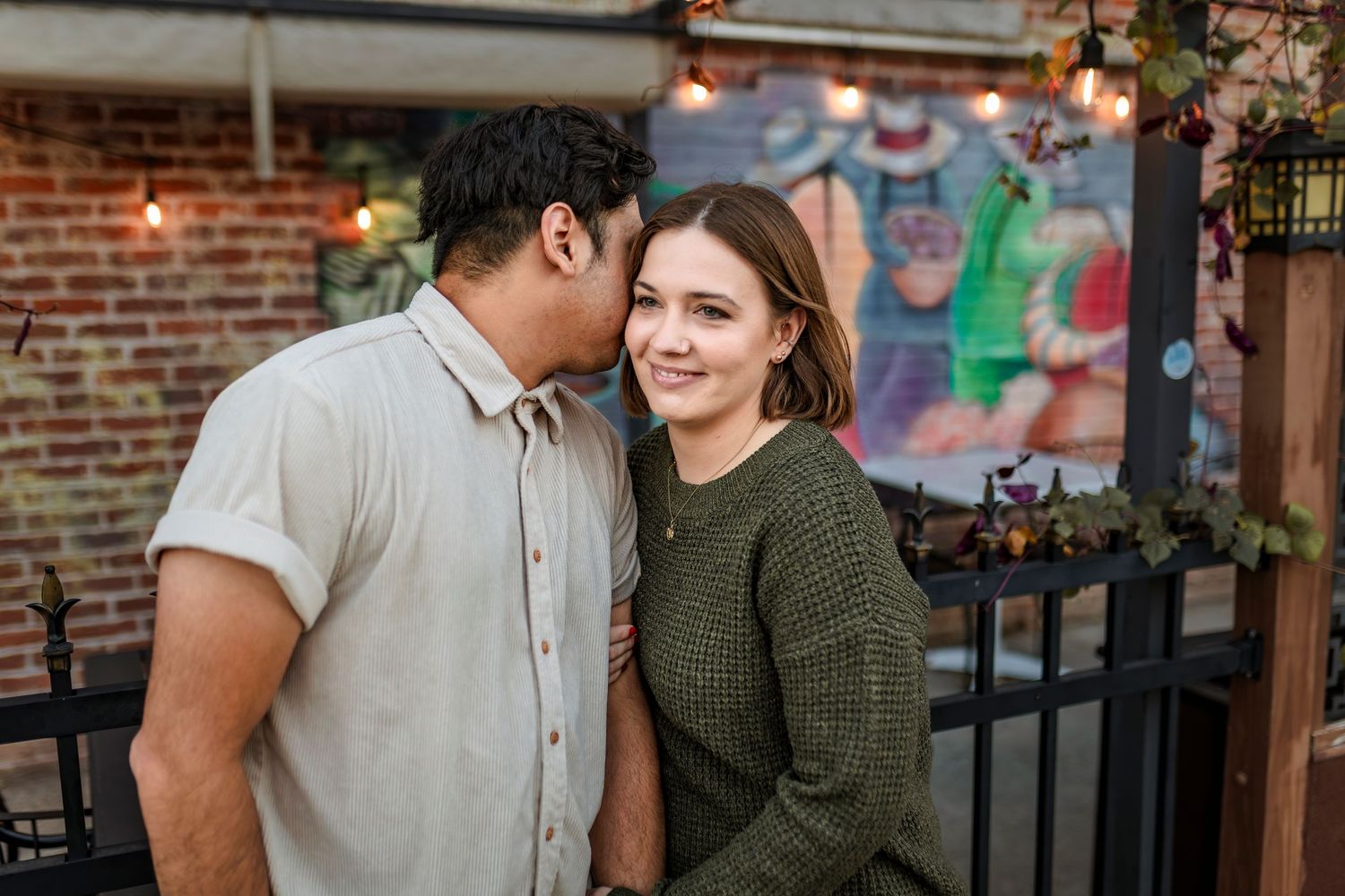 A romantic couple shares an intimate moment on a brick patio under twinkling string lights.