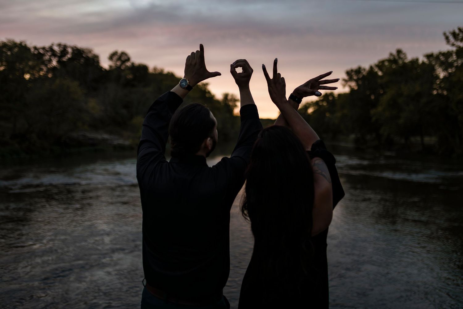 Silhouetted figures make peace signs against a moody sunset sky by a lake.