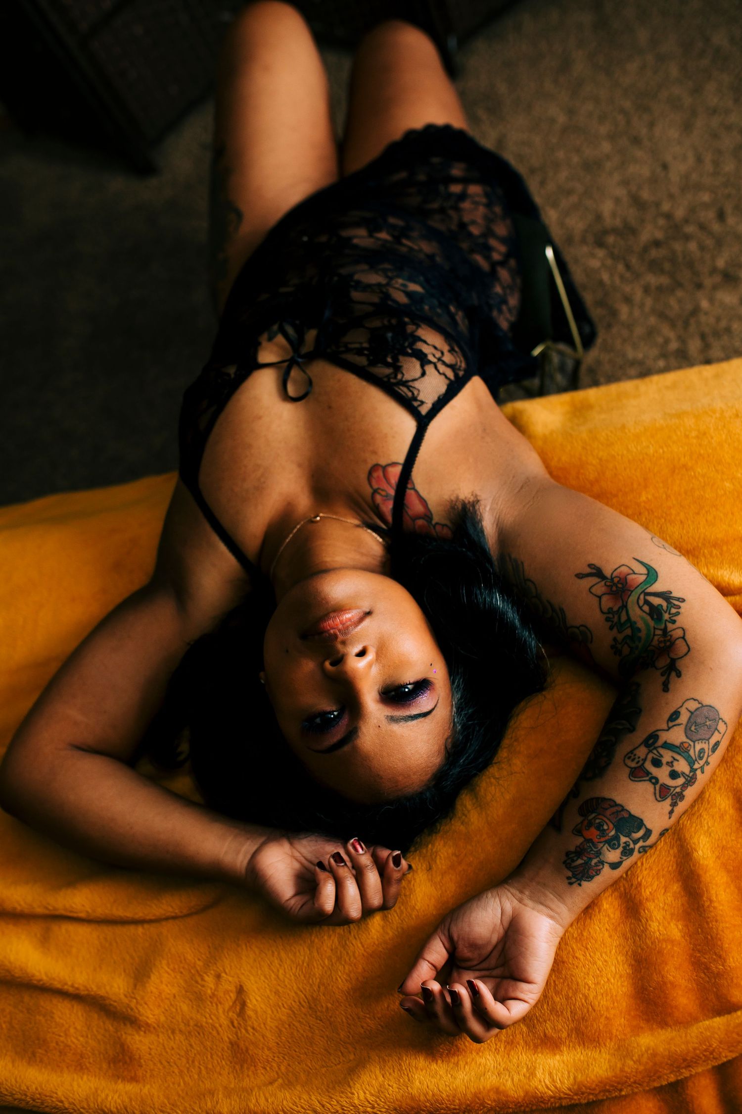 Woman in black lace top laying on orange blanket showing artistic arm tattoos in moody boudoir photo.