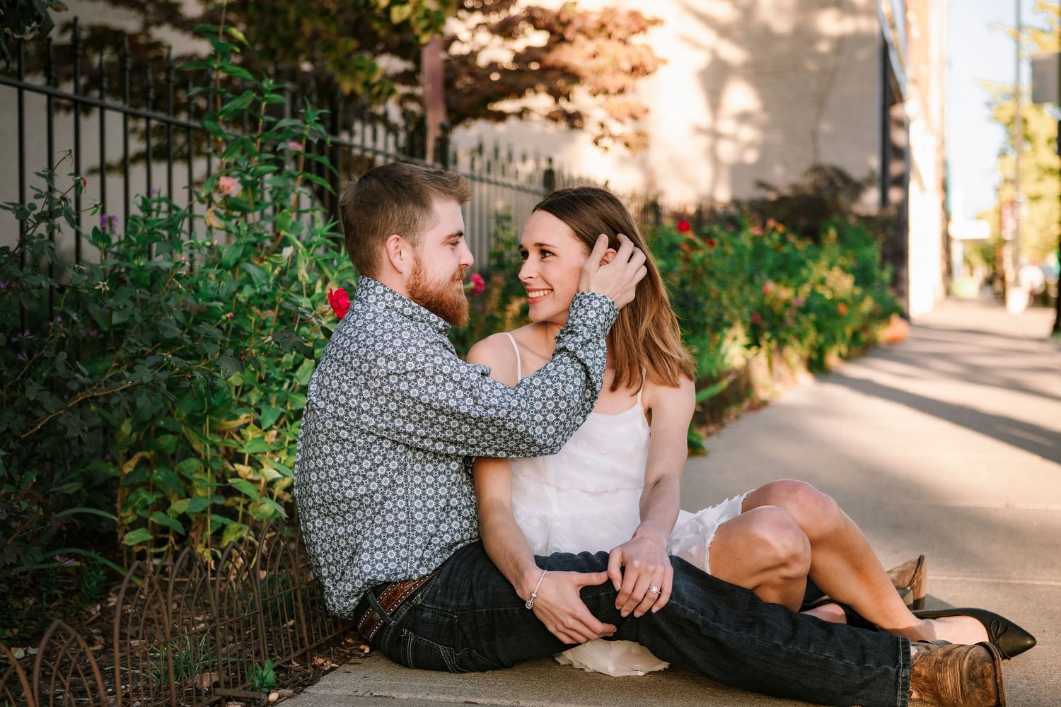 A couple sitting together on a sunlit sidewalk next to flowering bushes, sharing an intimate moment in summer.