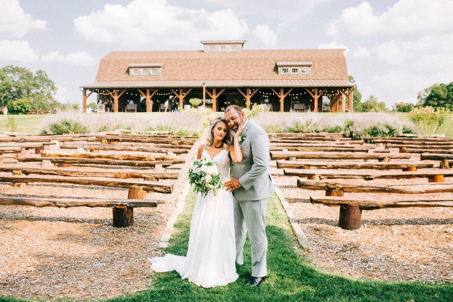 Bride and groom embrace at rustic outdoor venue with wooden bench seating.