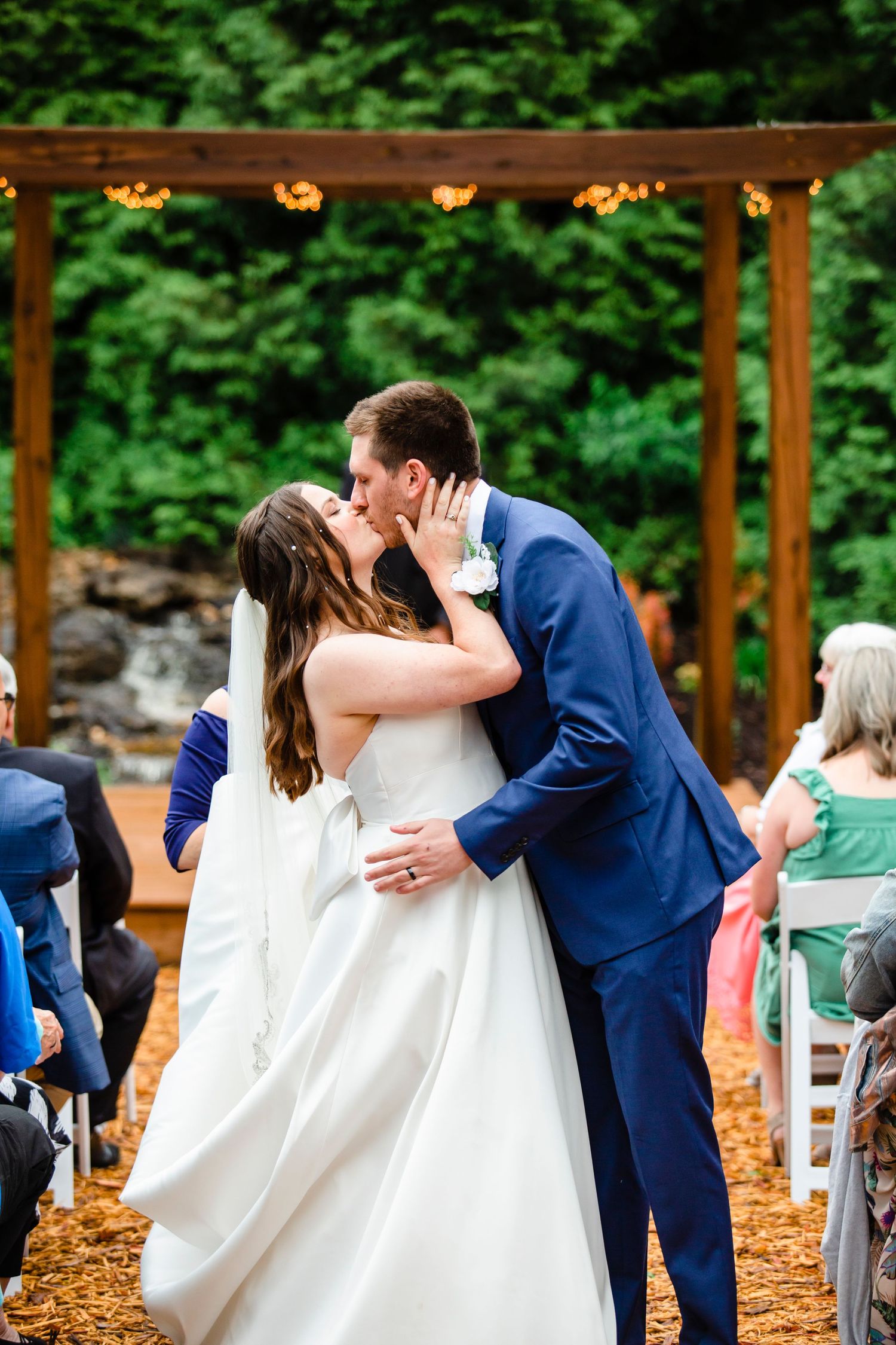 Wedding celebration at an outdoor venue with wooden pergola and white chairs.