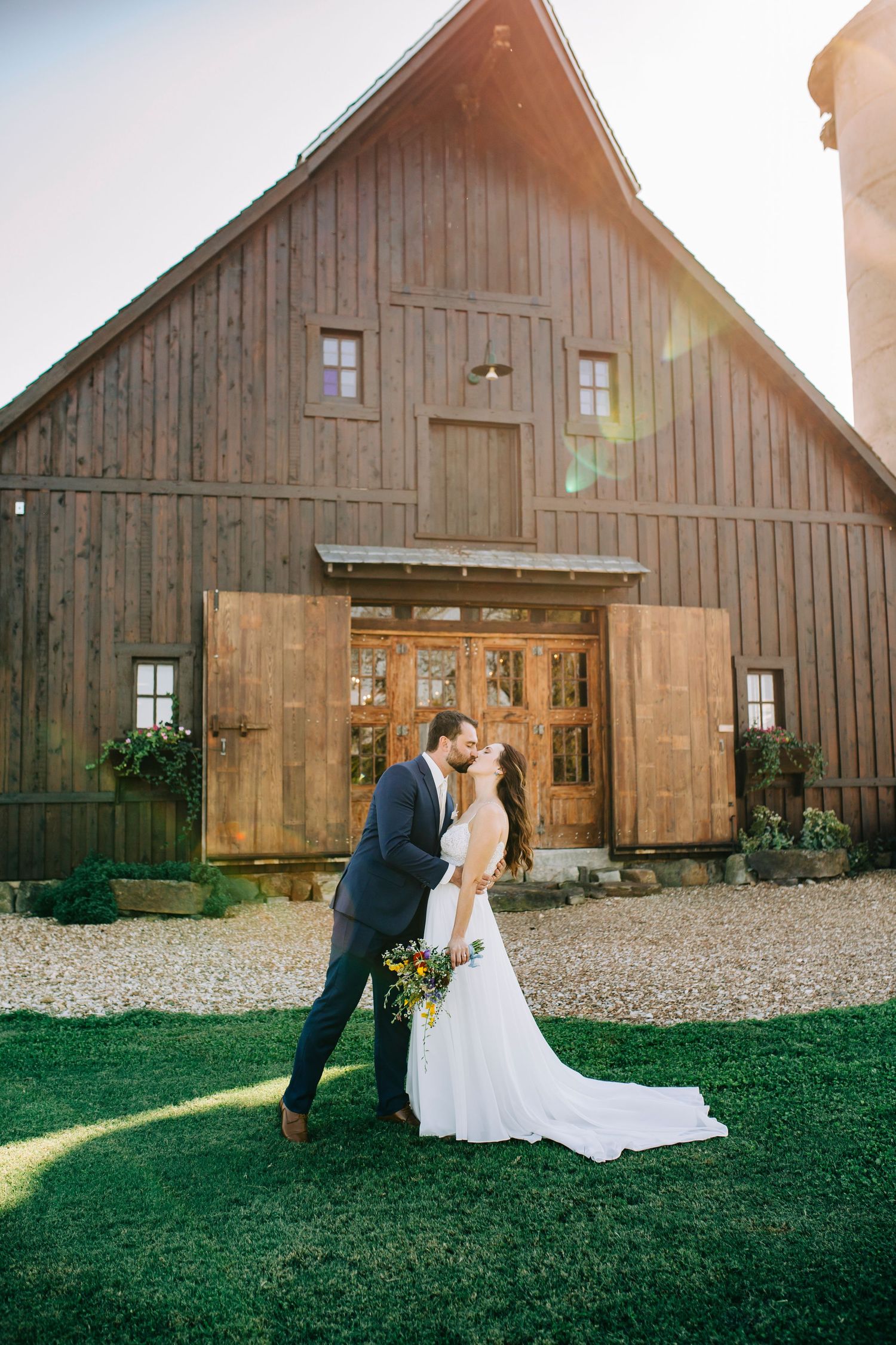 Couple shares romantic moment in front of rustic barn wedding venue with wooden doors and green lawn.