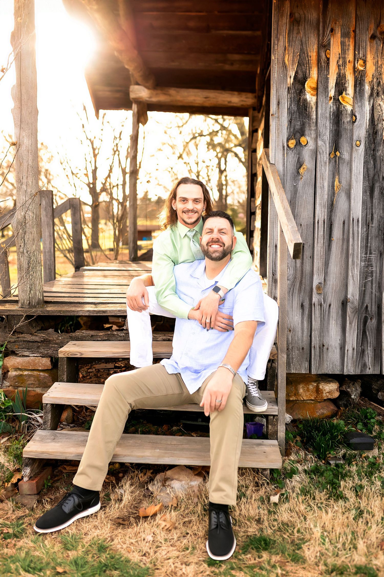 Two people in casual attire sitting together on wooden steps outside a rustic cabin with warm sunlight in background.