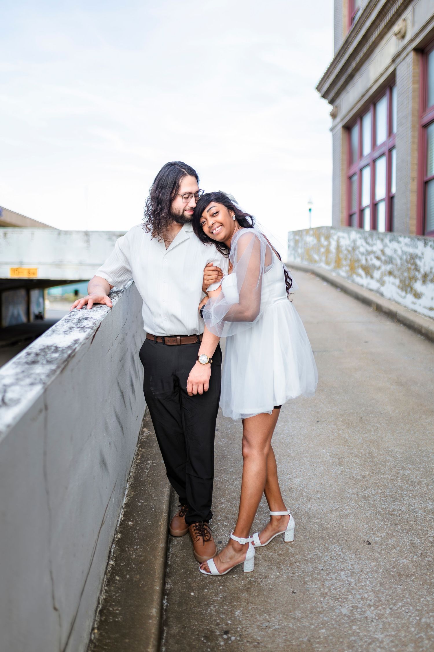 A couple in white and black attire share a romantic moment while leaning against a concrete railing on an urban walkway.