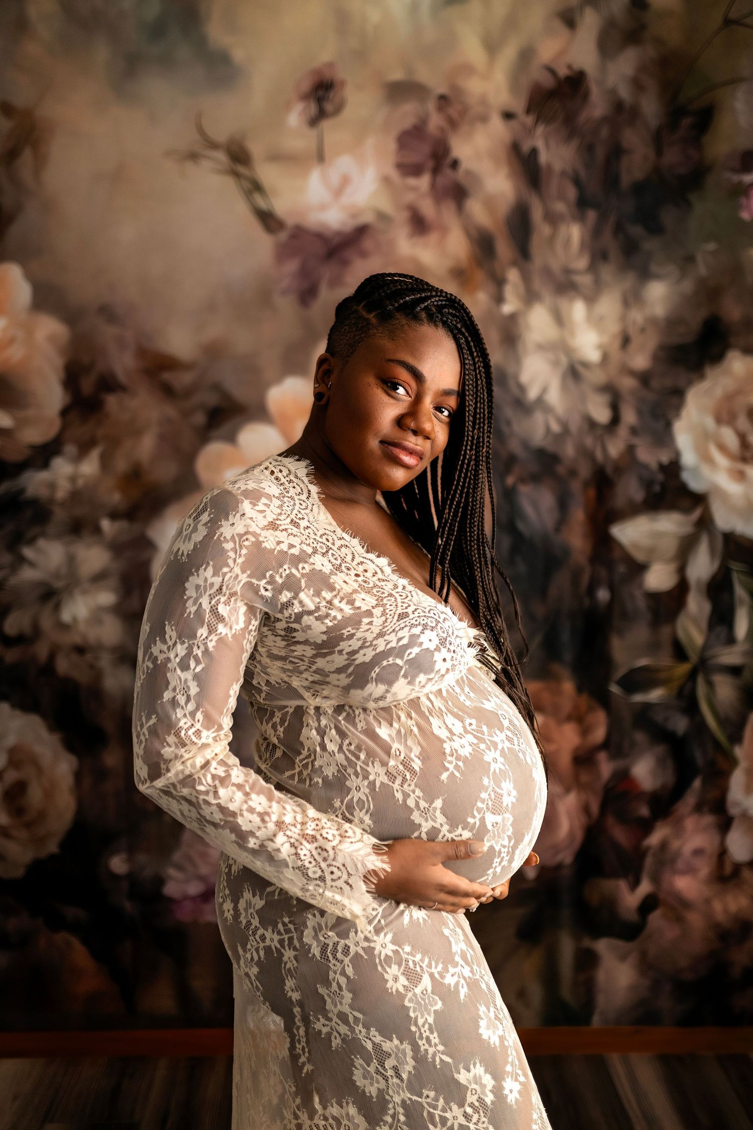 Expecting mother poses in white lace maternity gown against moody floral backdrop in warm lighting.