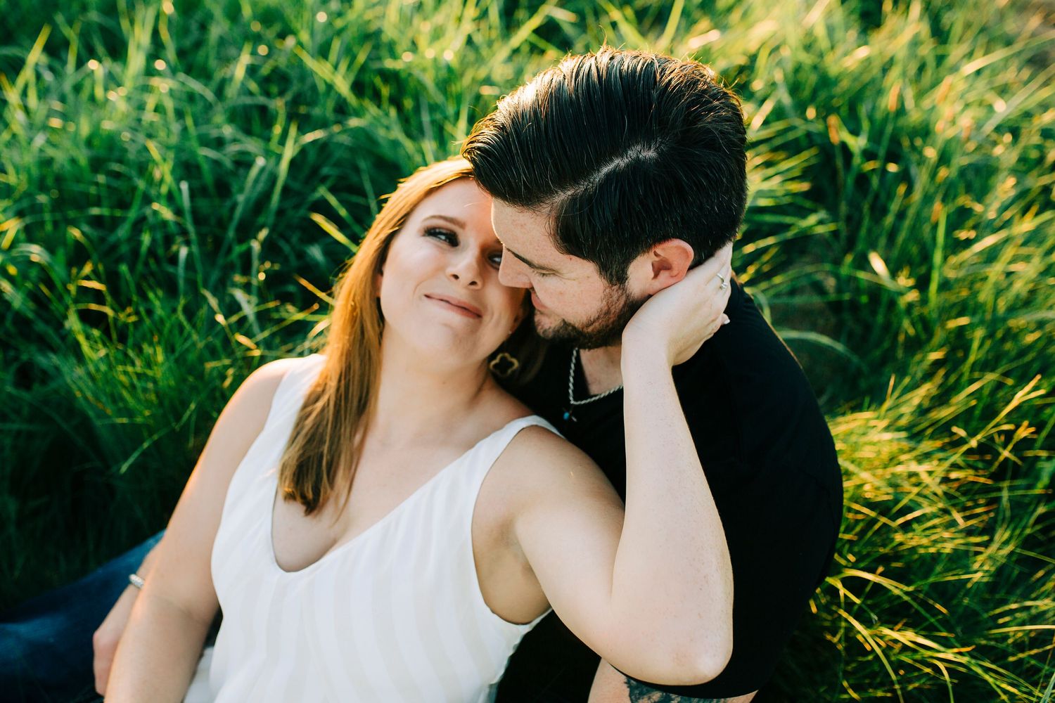 A couple sitting together in tall green grass on a sunny afternoon, sharing an intimate moment.