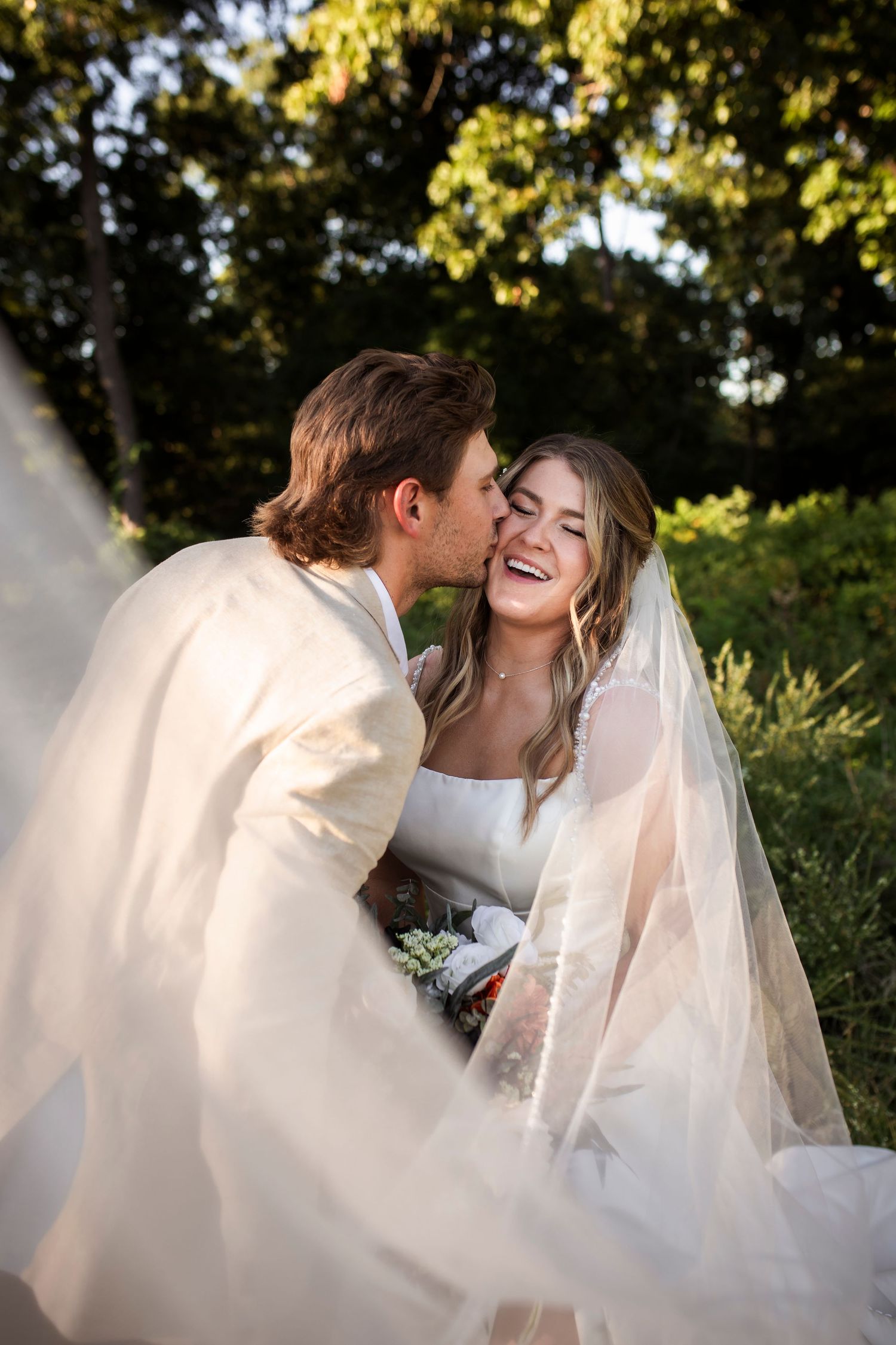 Joyful wedding couple shares a tender moment outdoors with sunlit trees in the background and flowing veil in foreground.