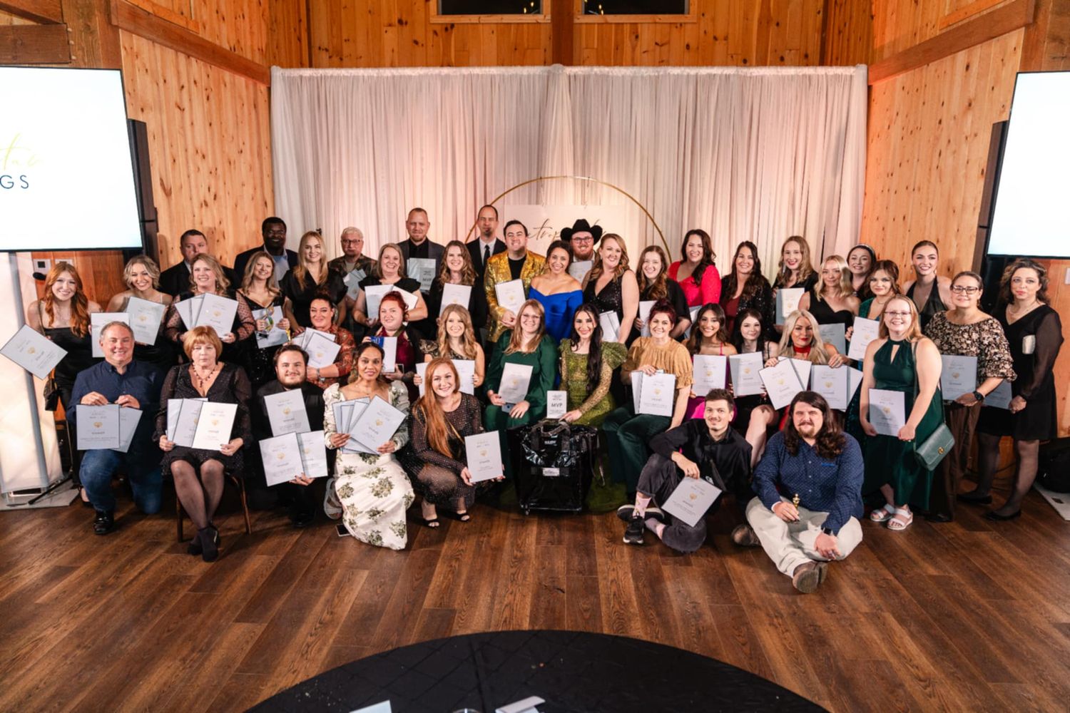 Large group of people holding certificates pose together on wooden floor in front of white backdrop at awards ceremony.