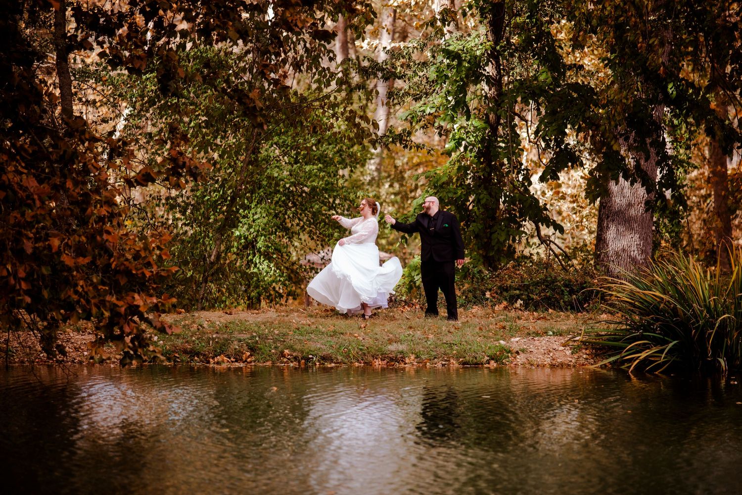 A couple in formal attire poses together on a wooden bridge surrounded by autumn foliage in a romantic outdoor setting.