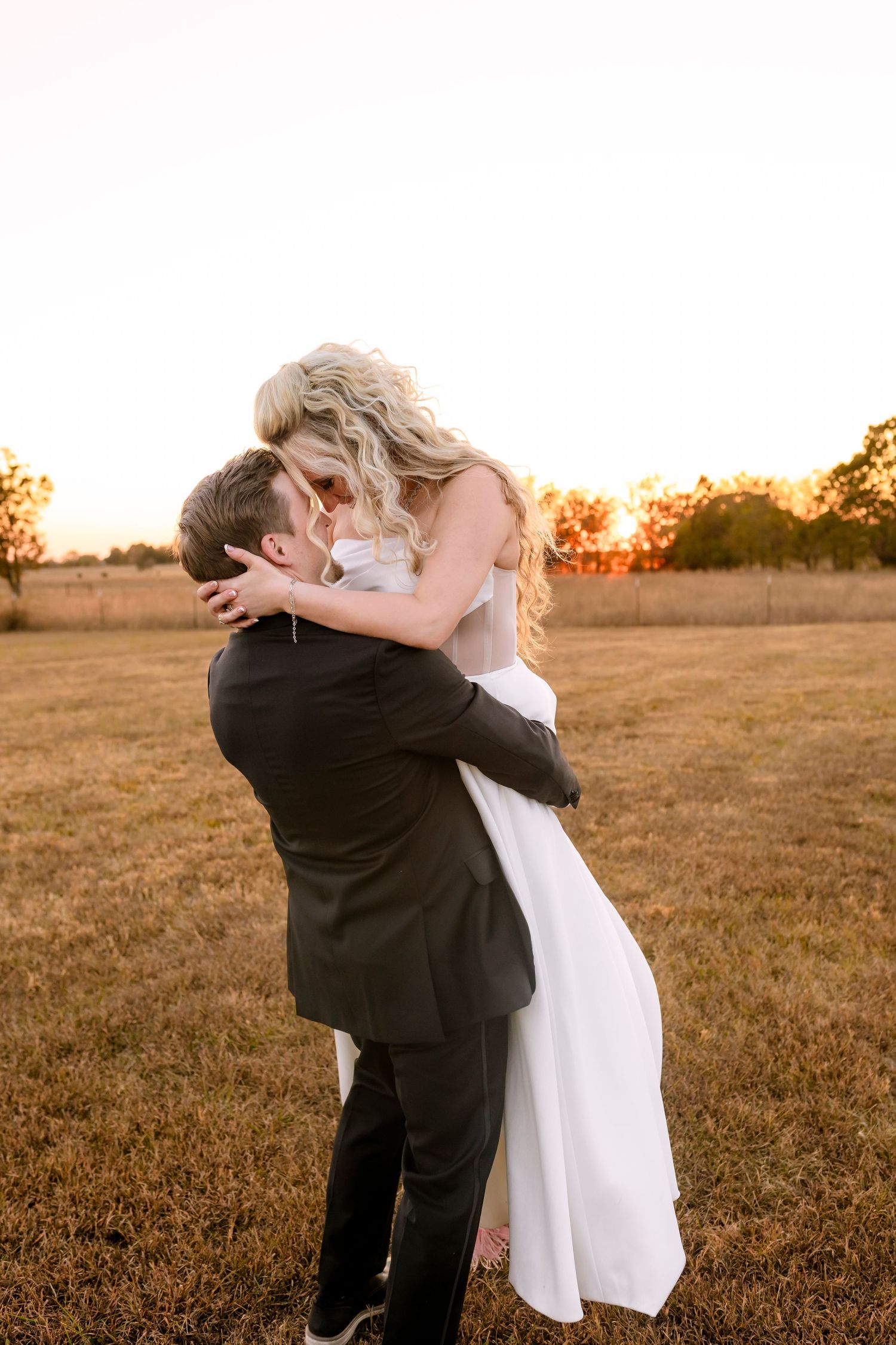 A couple embraces in a romantic kiss during golden hour in an open field at sunset.