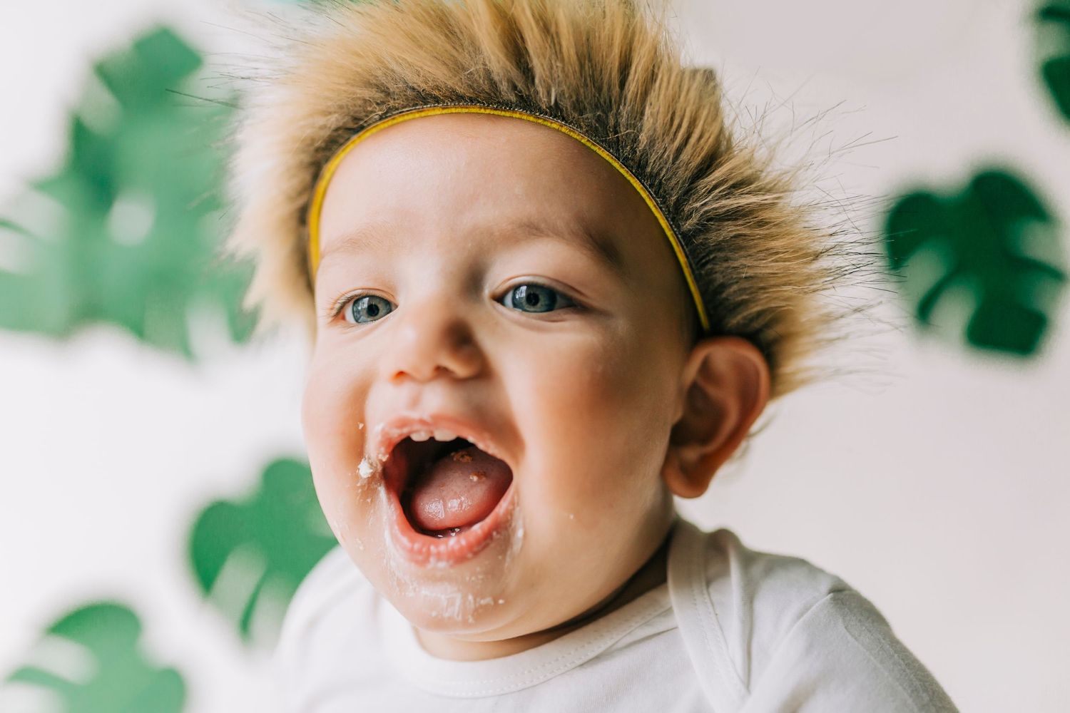 Laughing baby with spiky hairstyle poses against white backdrop with green monstera leaf decorations in background.