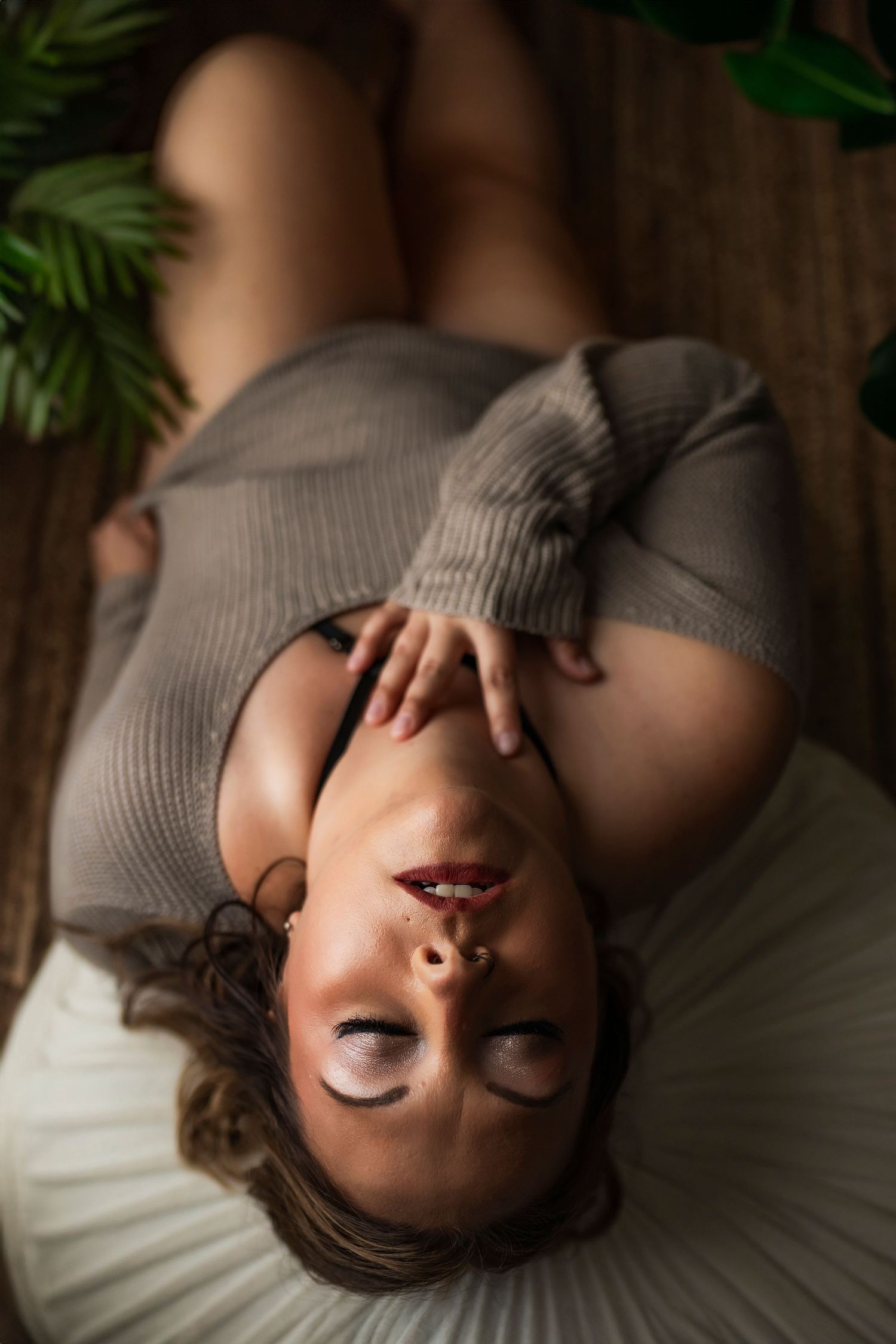 A woman wearing a grey ribbed sweater lies back on white bedding with houseplants visible in the soft background.
