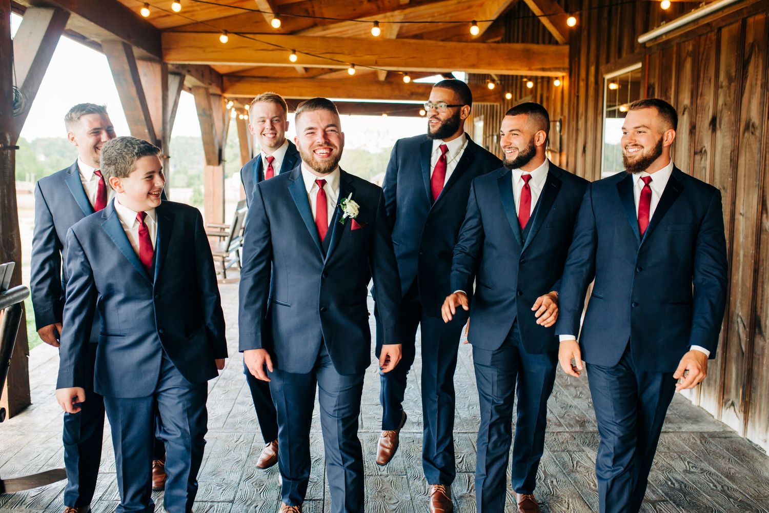 Groomsmen wearing navy suits and red ties walk together in a rustic venue.