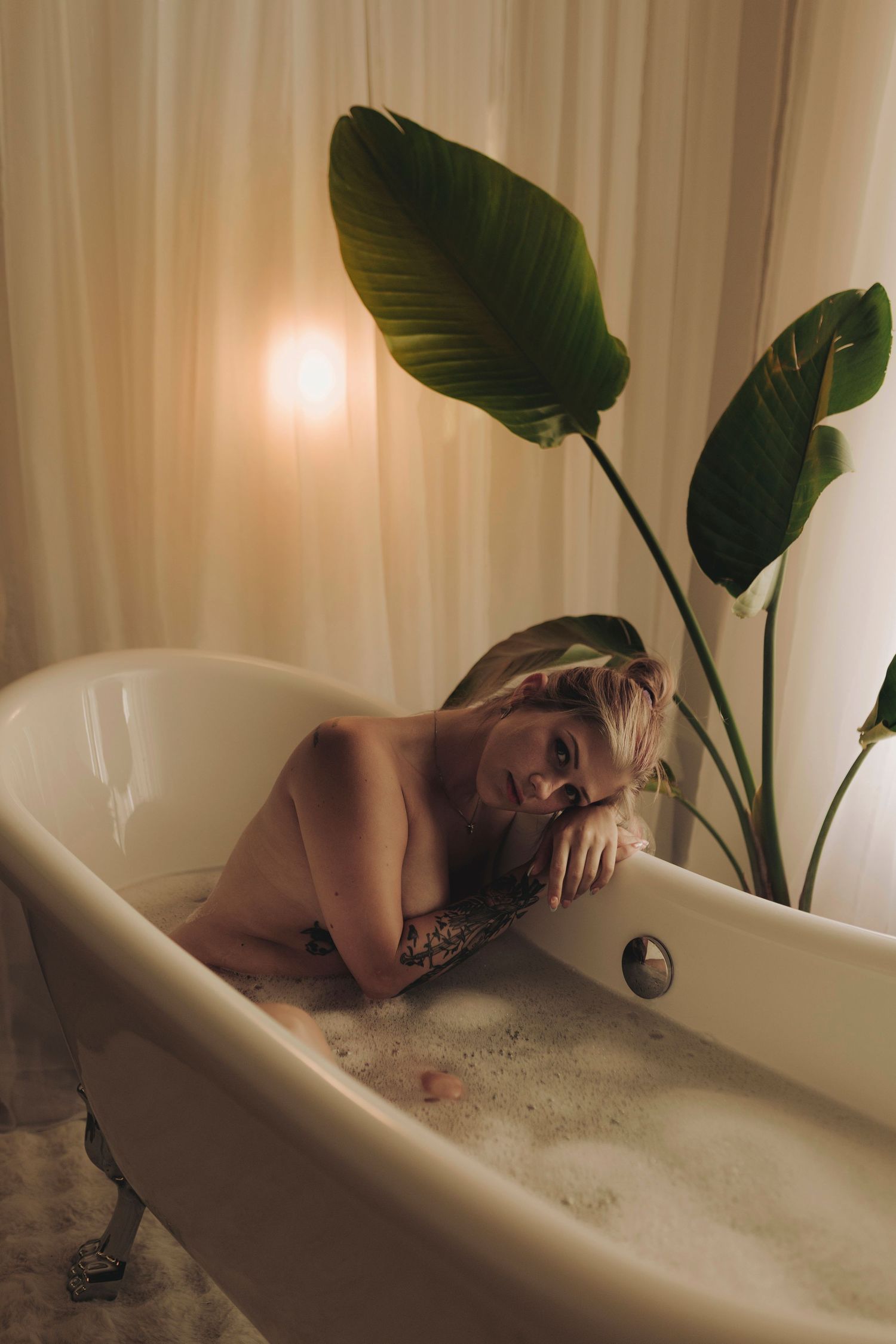Person relaxing in a bubble bath with tropical plants beside white freestanding bathtub in serene bathroom setting.