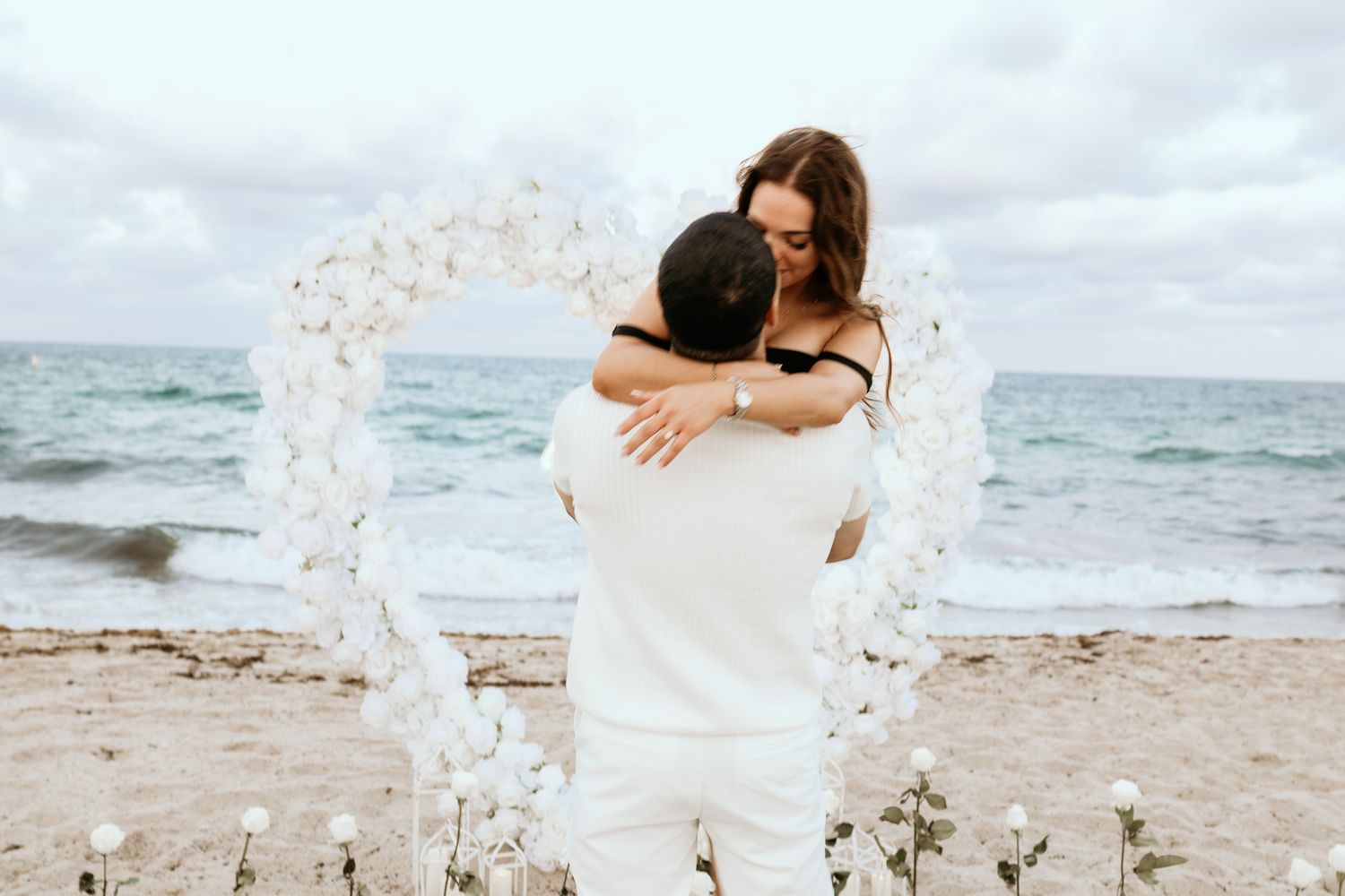 Couple embracing in front of a heart-shaped white floral arch on a sandy beach.
