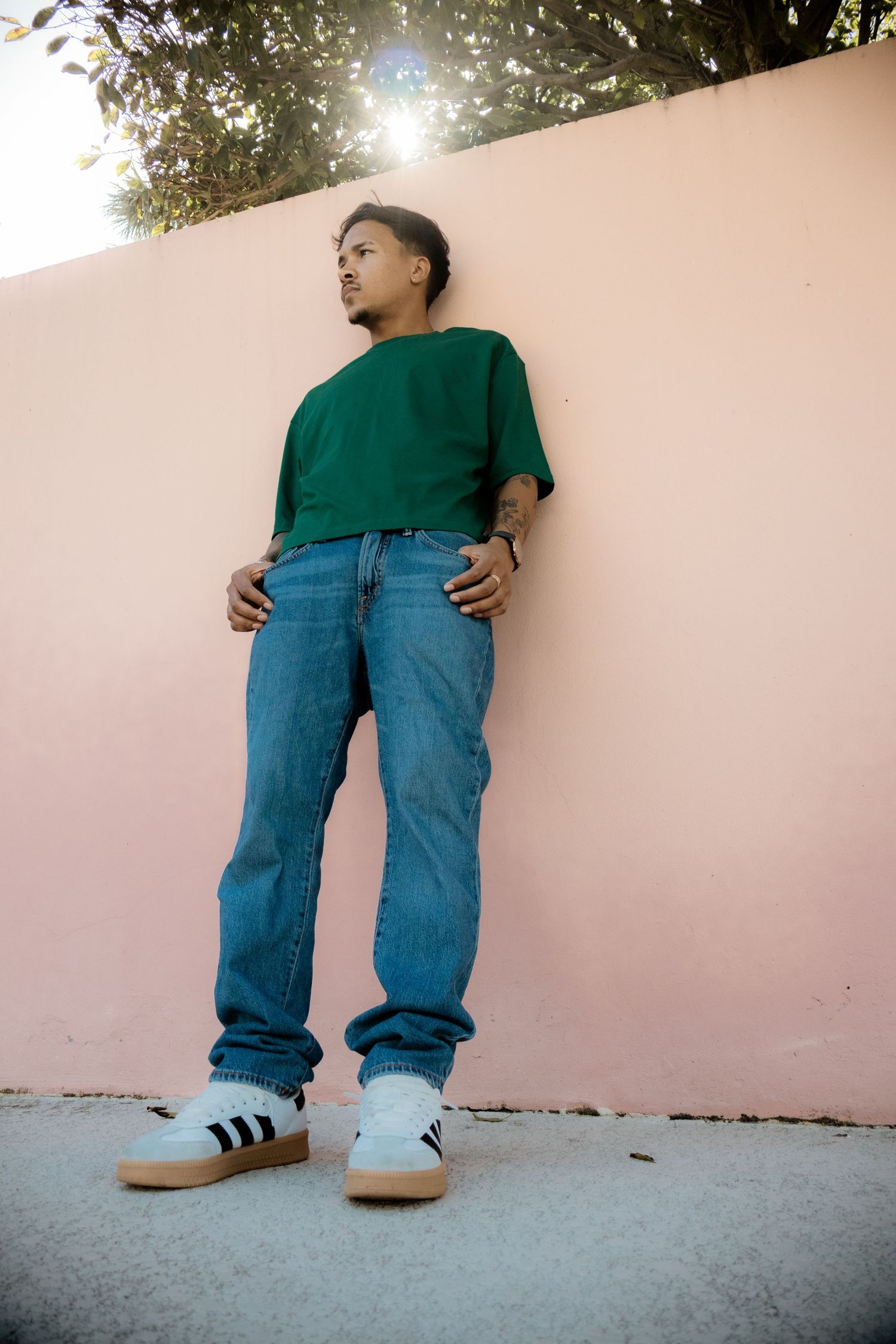 Young man in green tee and blue jeans leaning against a pink wall outdoors.