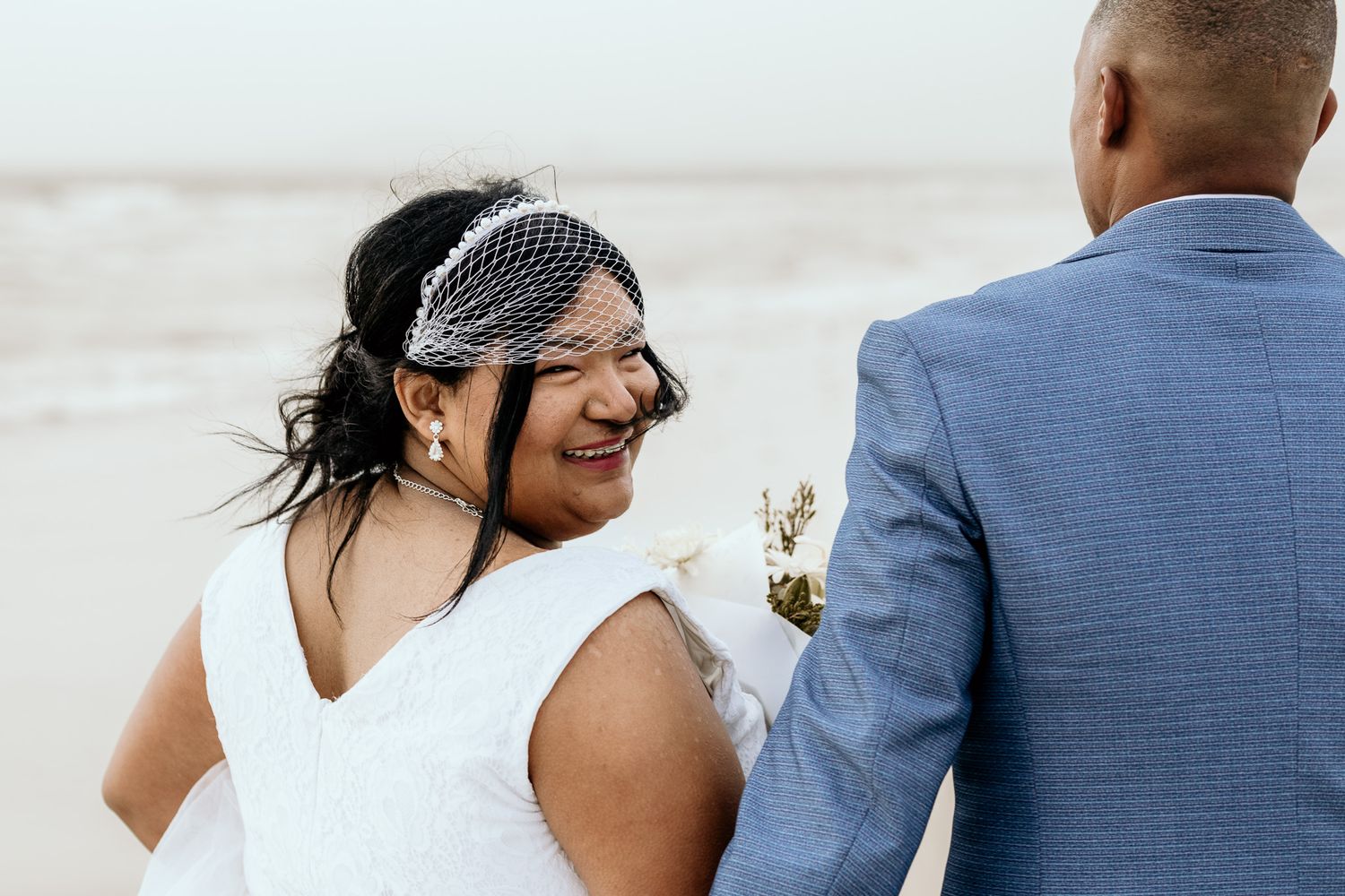 Radiant bride in birdcage veil smiling at groom in blue suit during their beach wedding celebration.