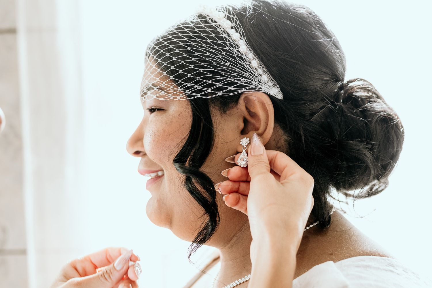 Smiling bride in birdcage veil having her earring placed while preparing for her wedding ceremony.
