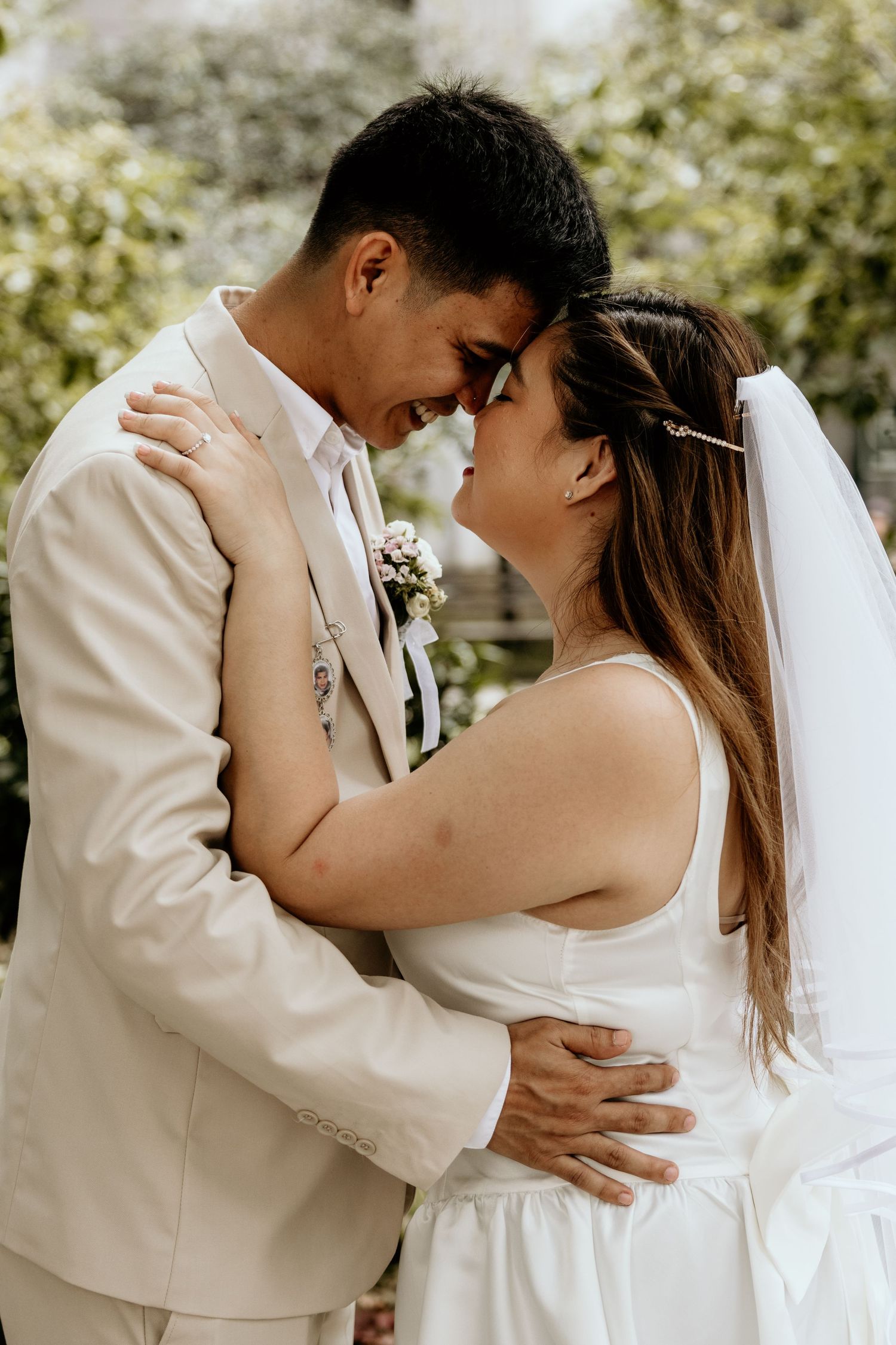 Newlywed couple in beige suit and white dress embrace forehead to forehead in garden.