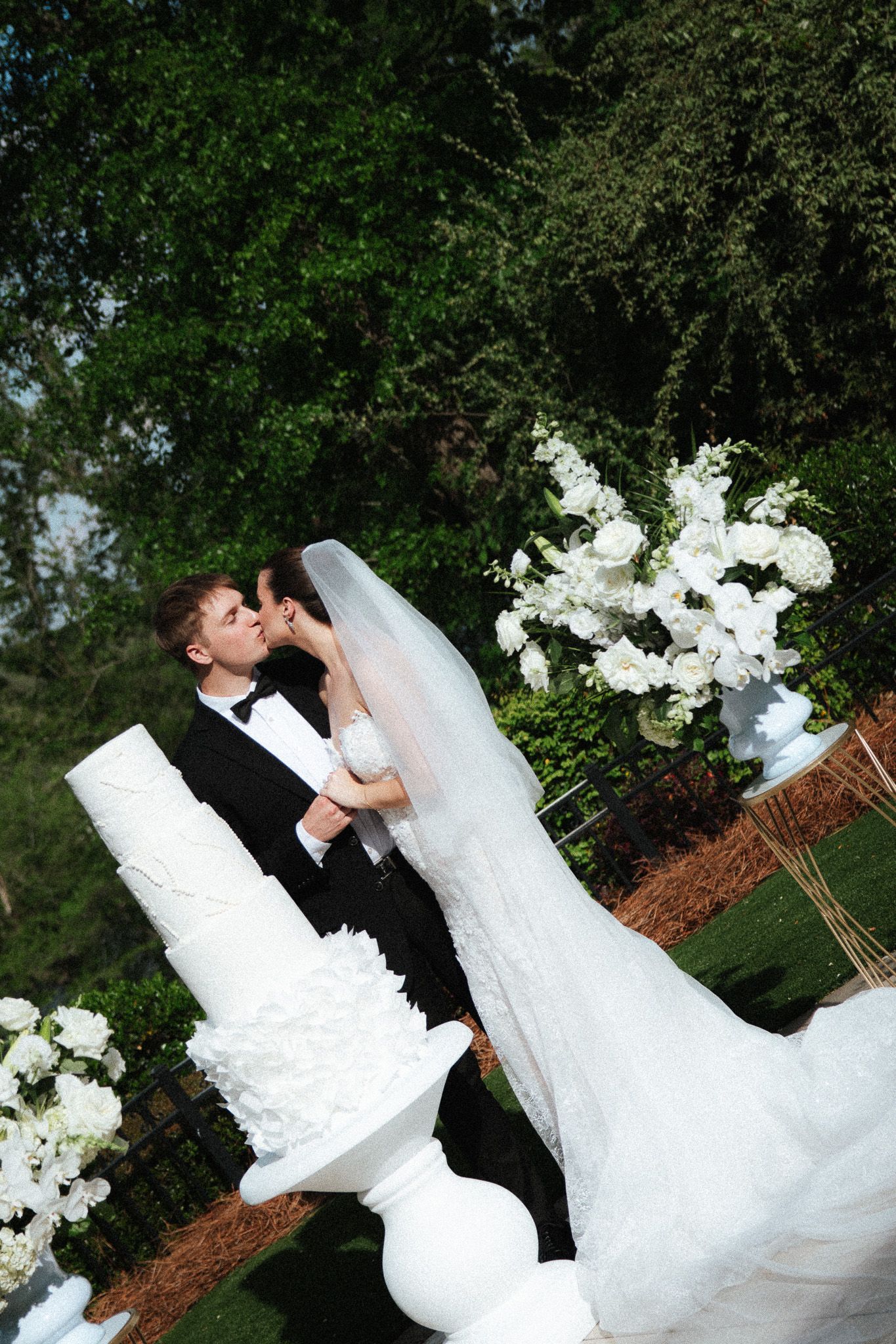 Bride and groom share a kiss at outdoor wedding ceremony with white floral arrangements and elegant cake.