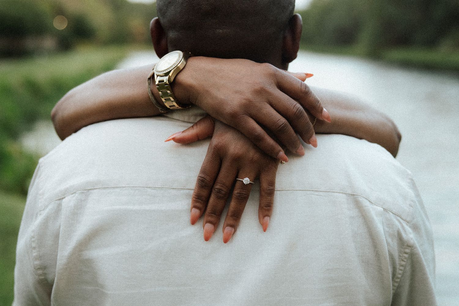 Close-up rear view of a woman's hands with engagement ring and bracelet resting on man's shoulders.