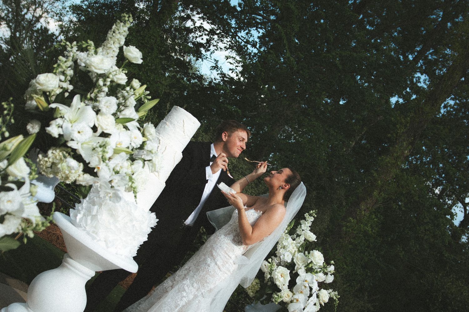 Bride and groom laughing together near a white wedding cake with lush floral arrangements in an outdoor setting.