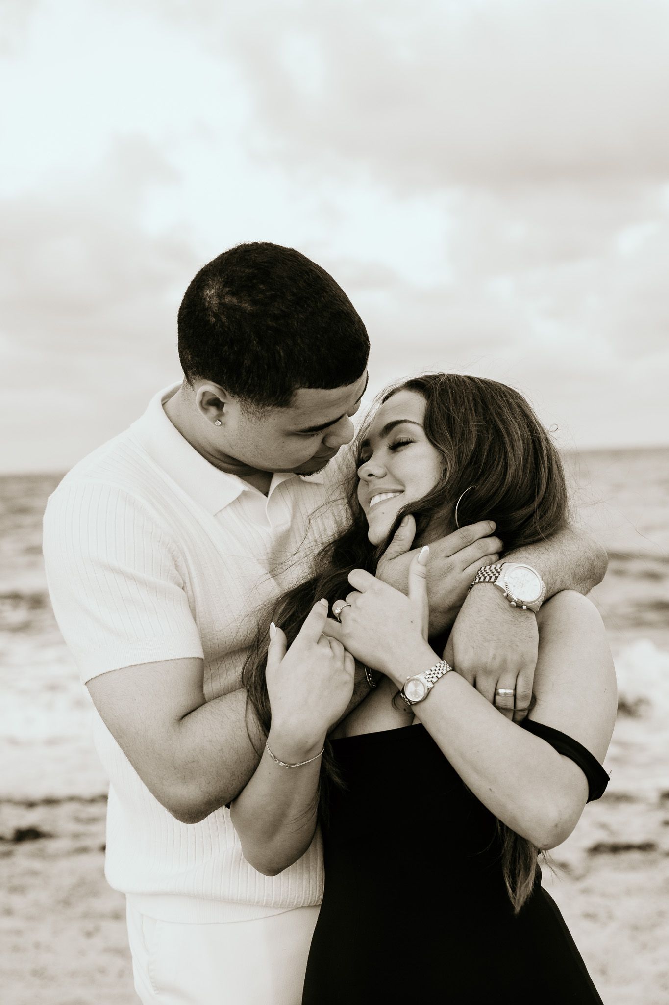 Black and white photo of a couple tenderly embracing on a breezy beach shoreline.