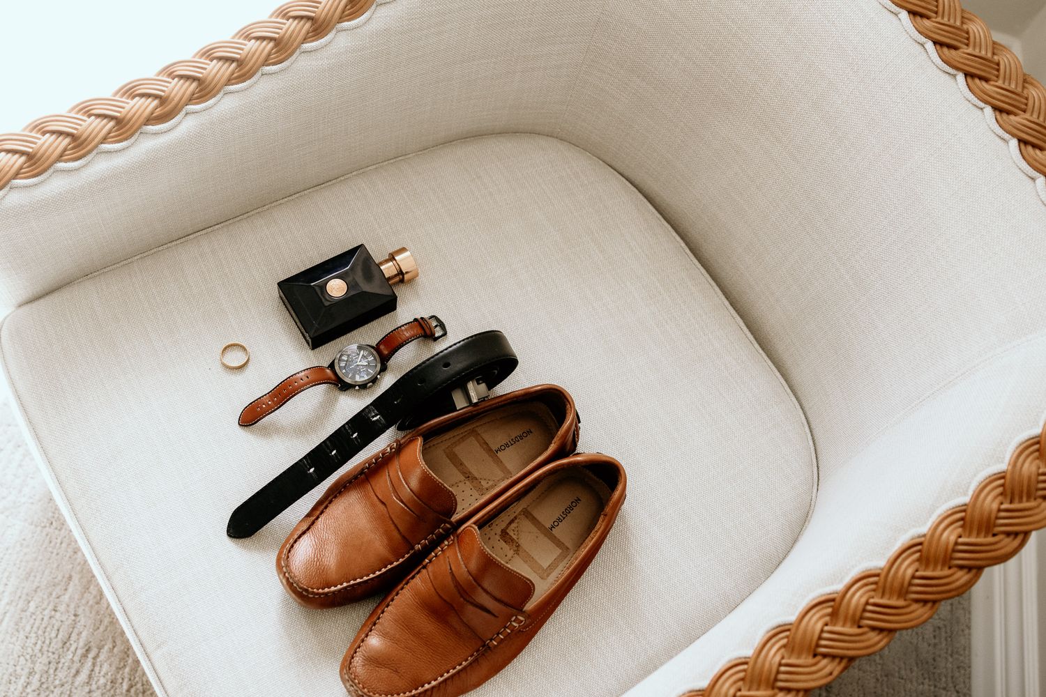 Groom accessories flat lay with brown loafers, belt, watch, ring, and cufflinks on woven chair.