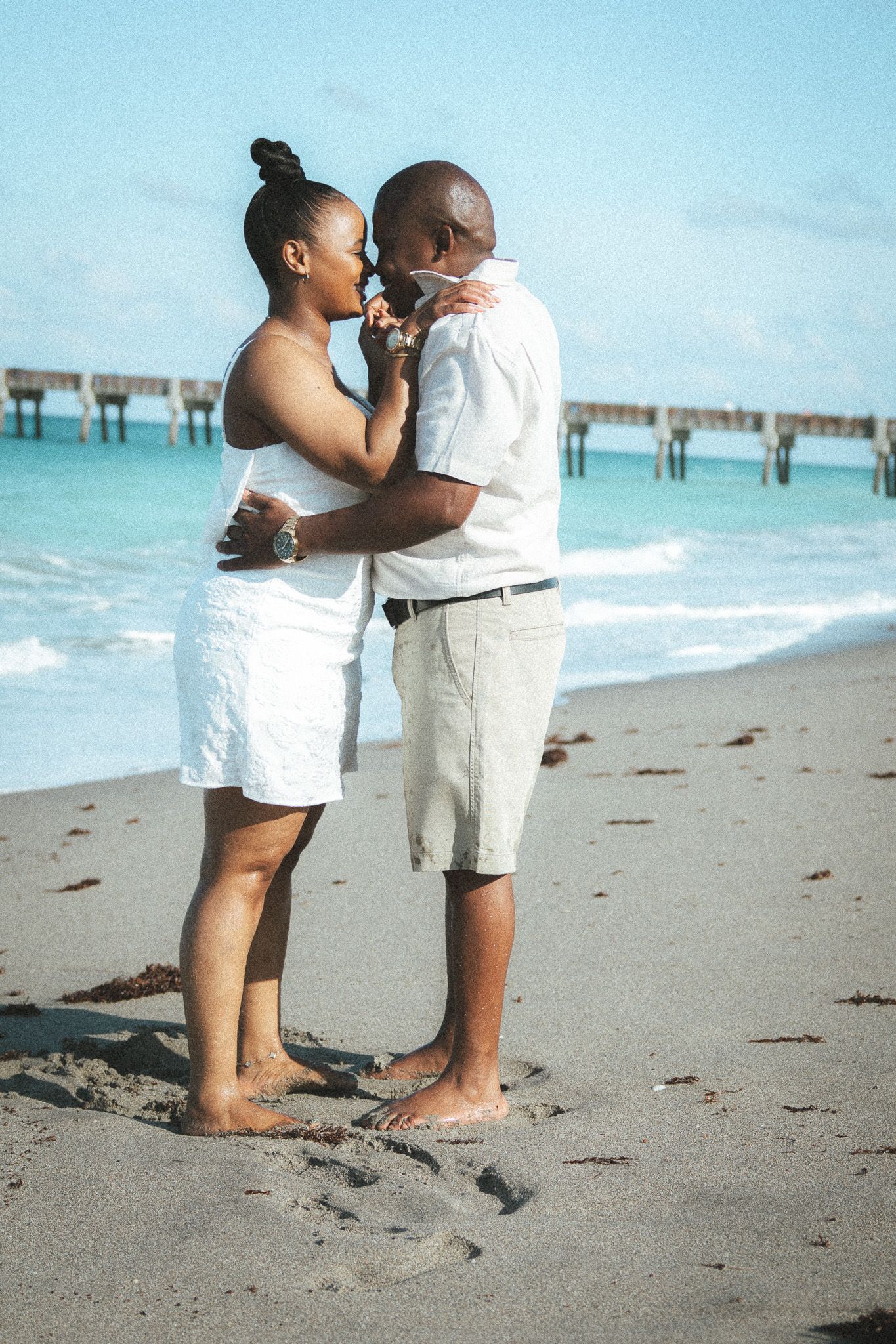 A couple in white outfits sharing a romantic moment on a sandy beach with turquoise ocean waves behind them.