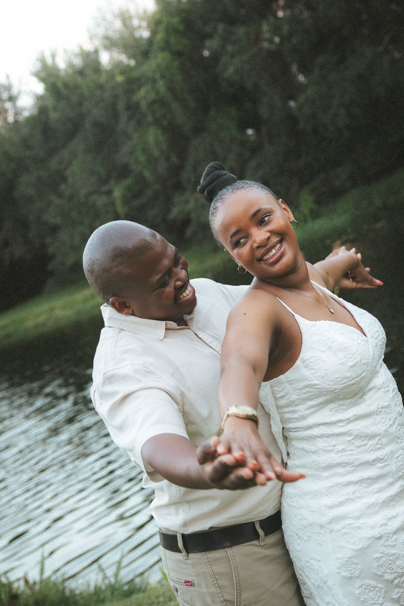 A happy couple dancing and laughing together outdoors near a pond surrounded by lush greenery.